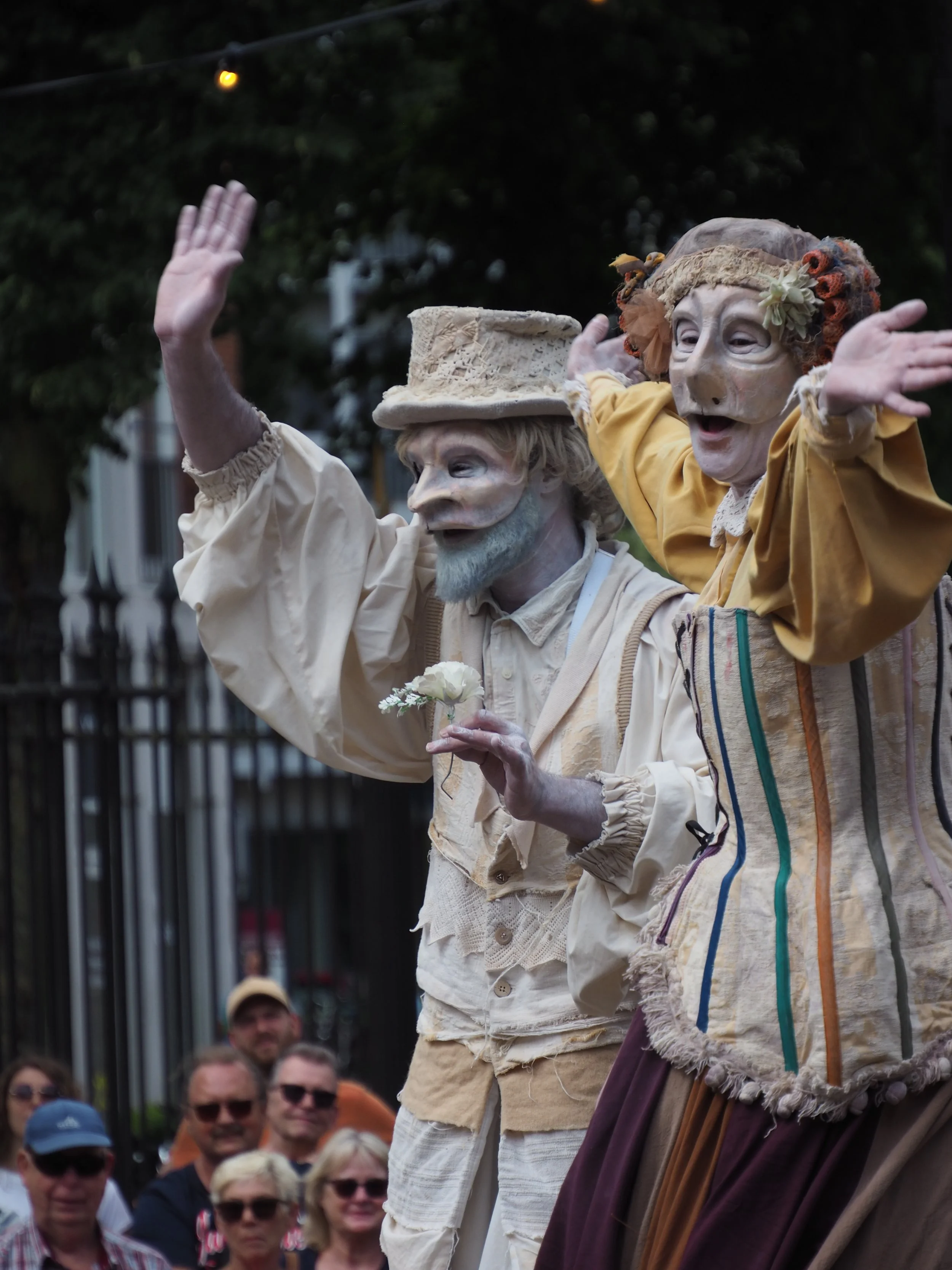 Two street performers dressed as puppet characters entertaining a crowd. The performers wear elaborate masks and costumes, and are engaging with the audience.