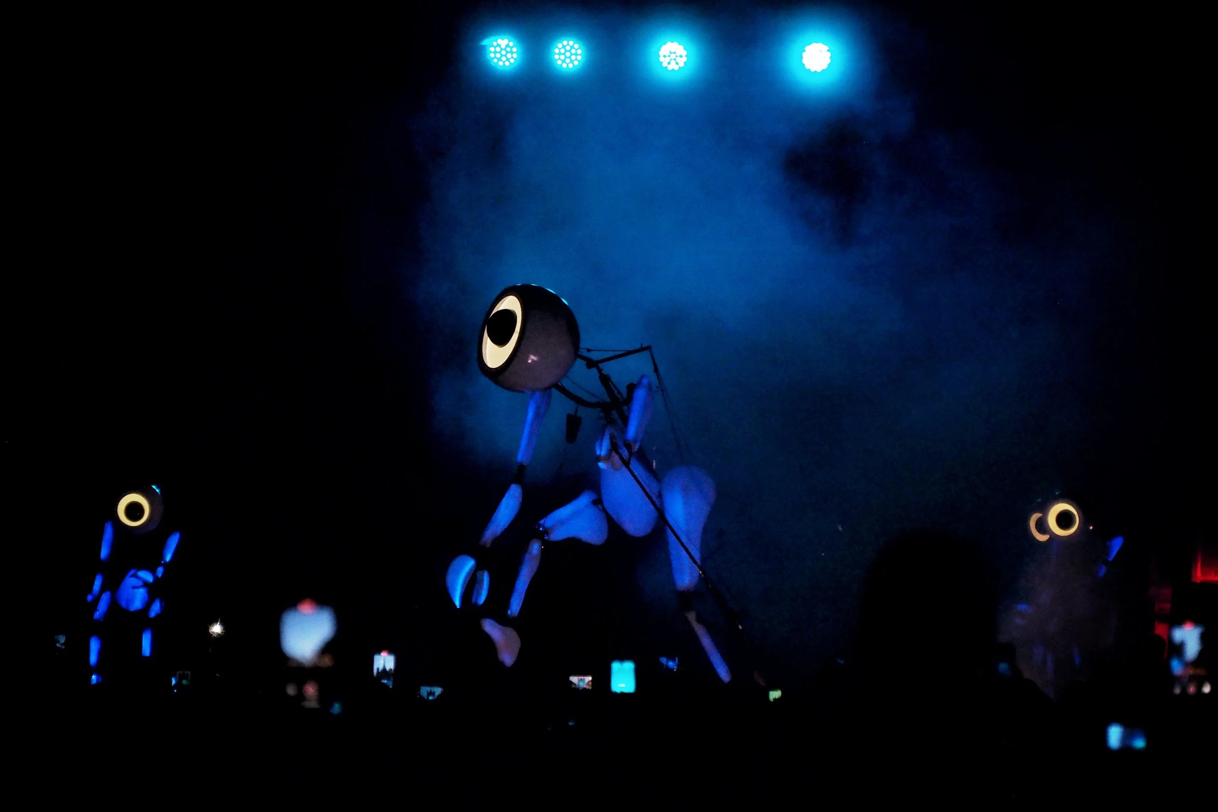 Night scene with a large mechanical robot with glowing eyes and blue lighting, surrounded by smaller figures or objects and illuminated by stage lights.