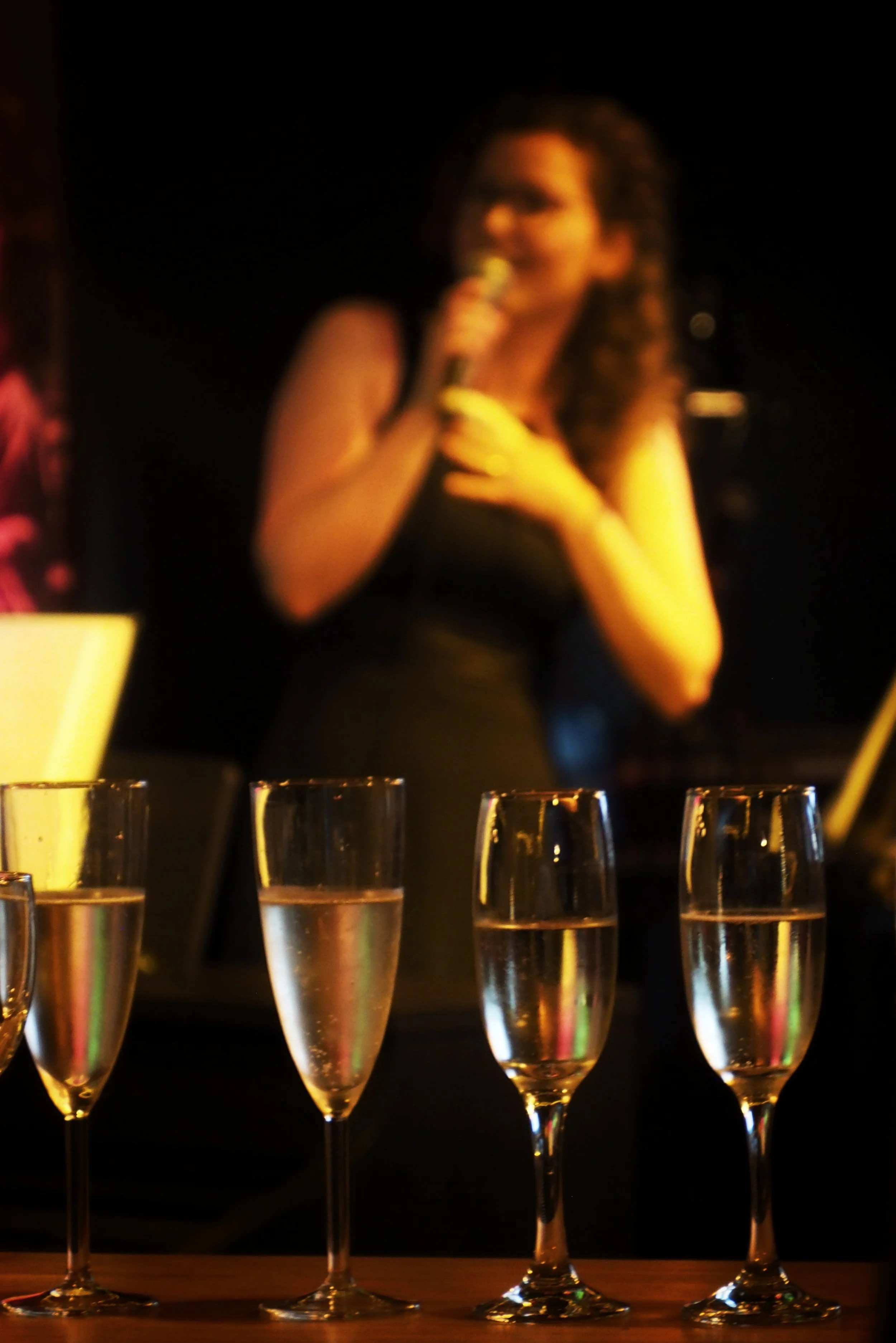 A woman with curly hair holding a microphone, standing behind a row of champagne glasses filled with a sparkling beverage, in a dark setting with a black background.