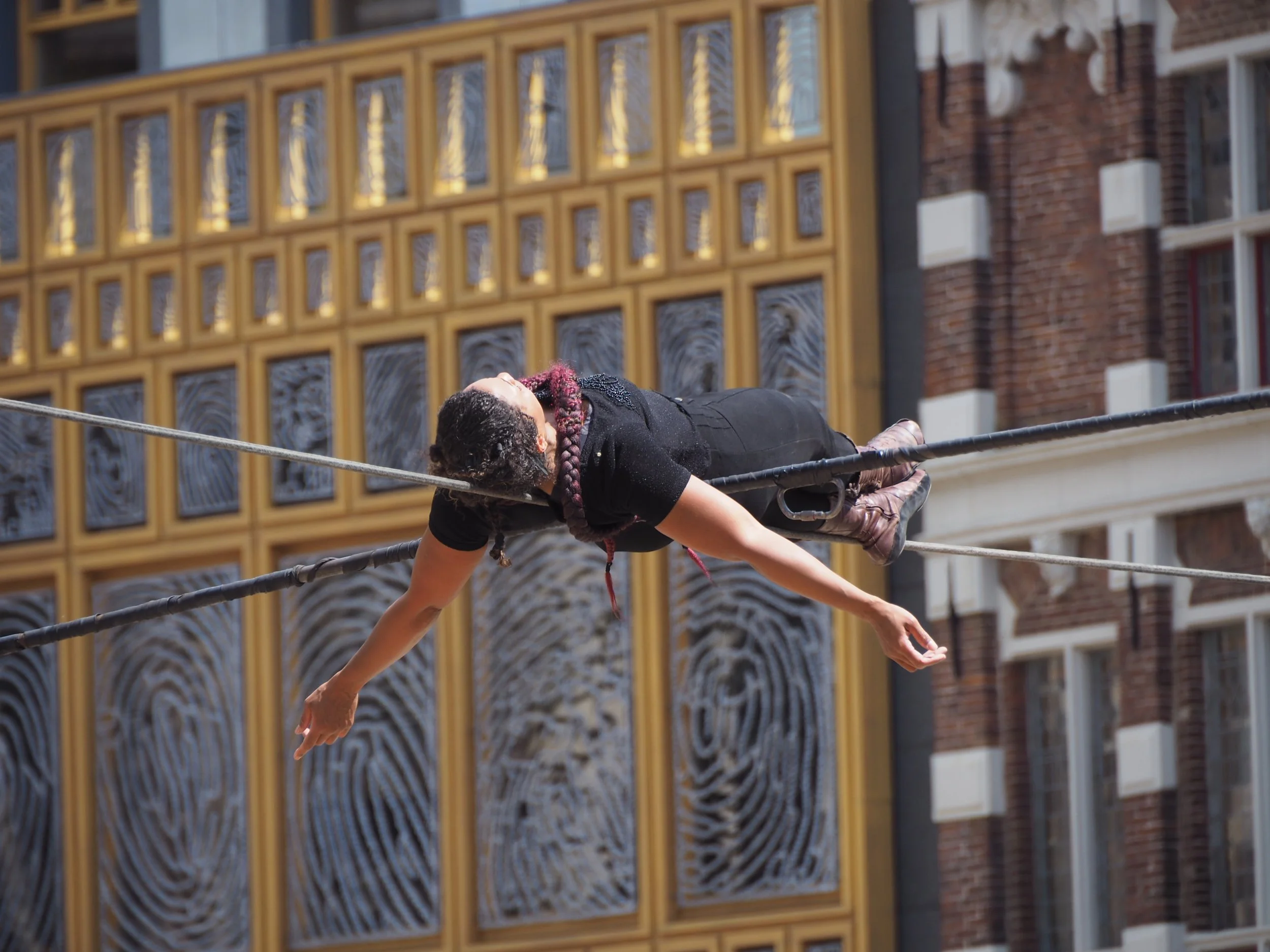 A person with long braided hair lying on a tightrope between buildings.