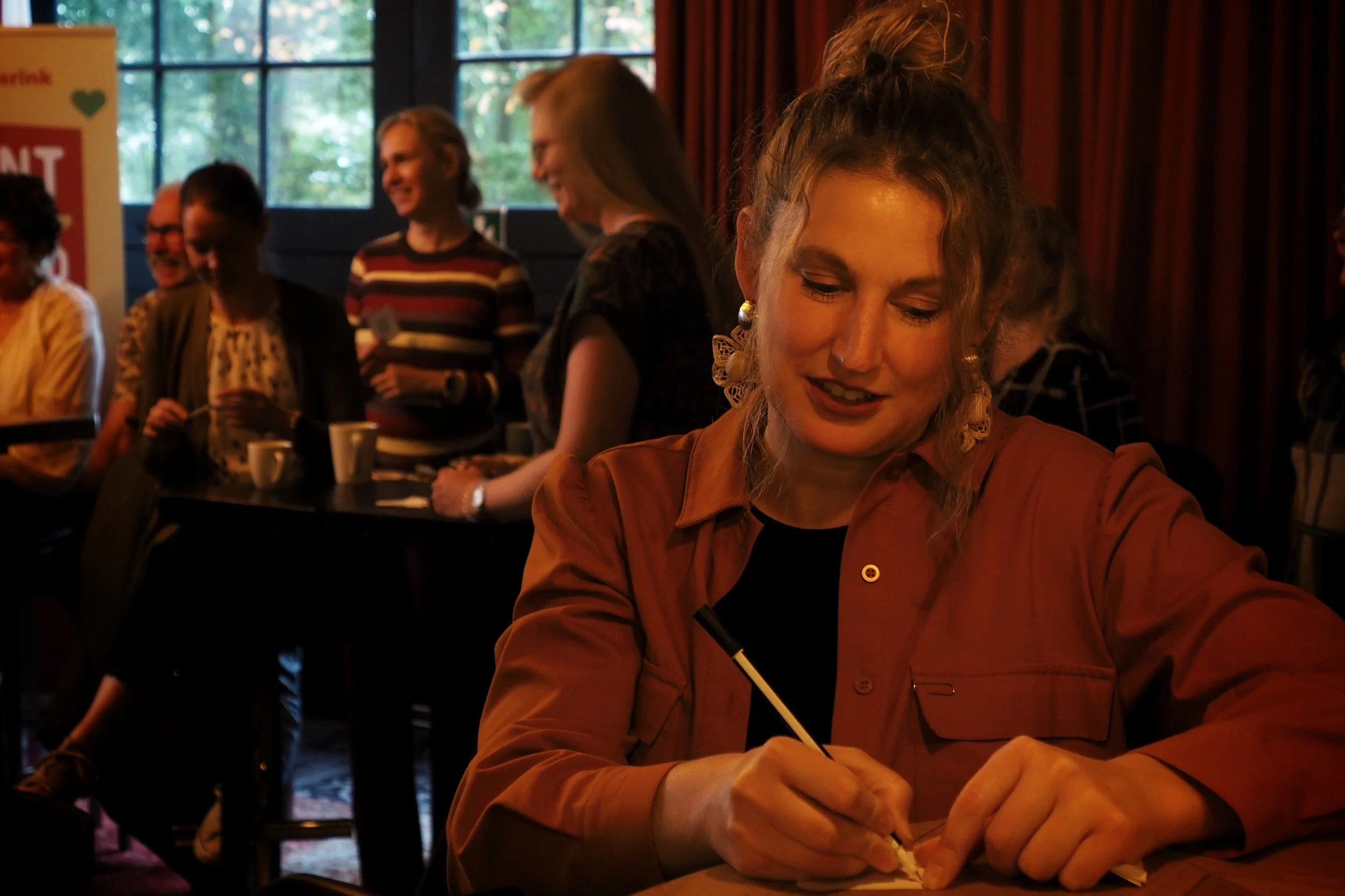 A woman with blonde curly hair tied up in a ponytail, wearing earrings and a burnt orange shirt, is sitting at a table writing with a black pen. In the background, there are several people standing and smiling, with large windows and red curtains beh