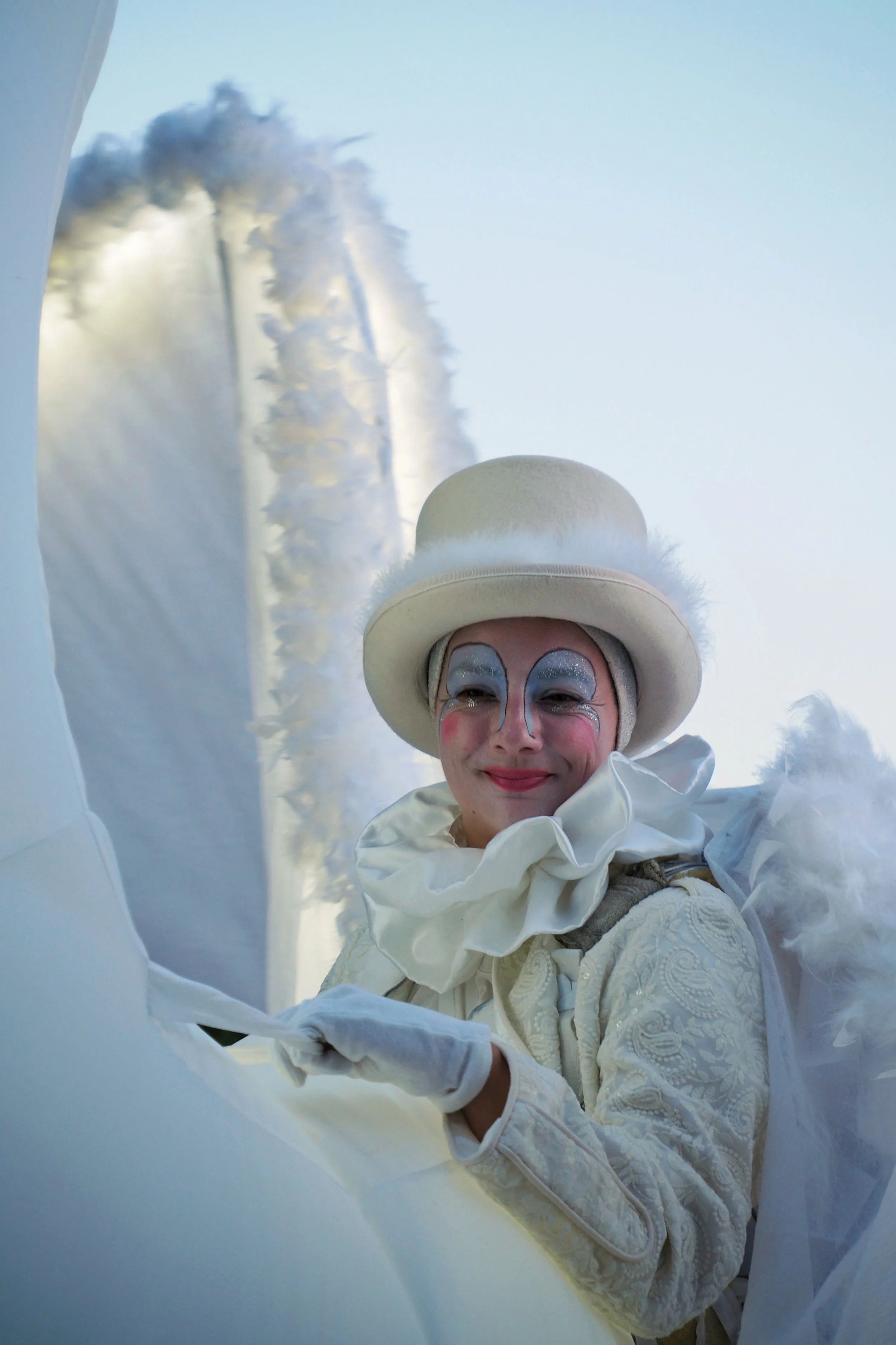 Close-up portrait of a joyful woman dressed in a white ornate outfit with a matching hat and silver makeup, smiling among white fabric and clouds in the background.