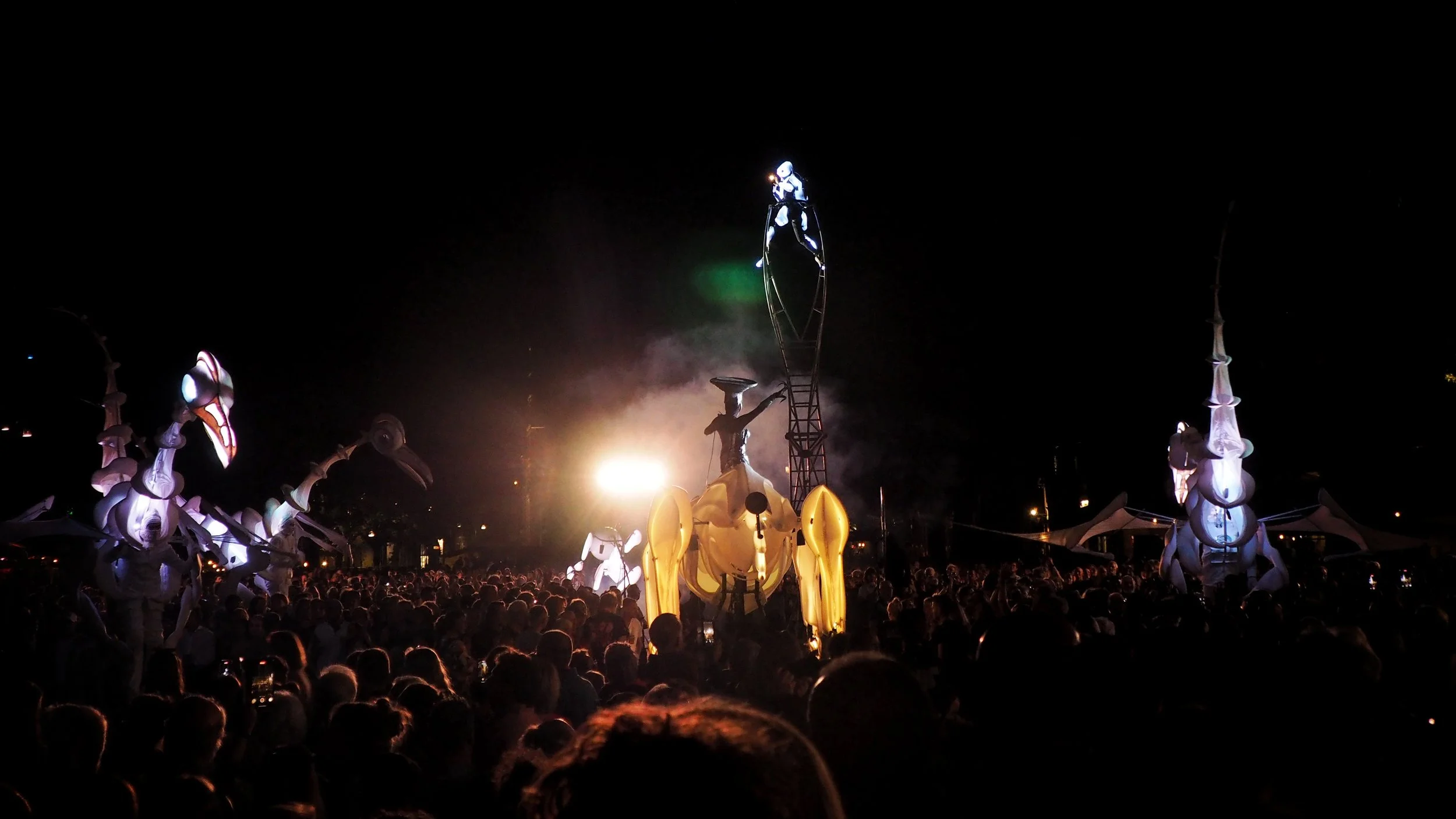 Nighttime scene of a large crowd watching a futuristic outdoor event with illuminated mechanical creatures and a performer on stilts.