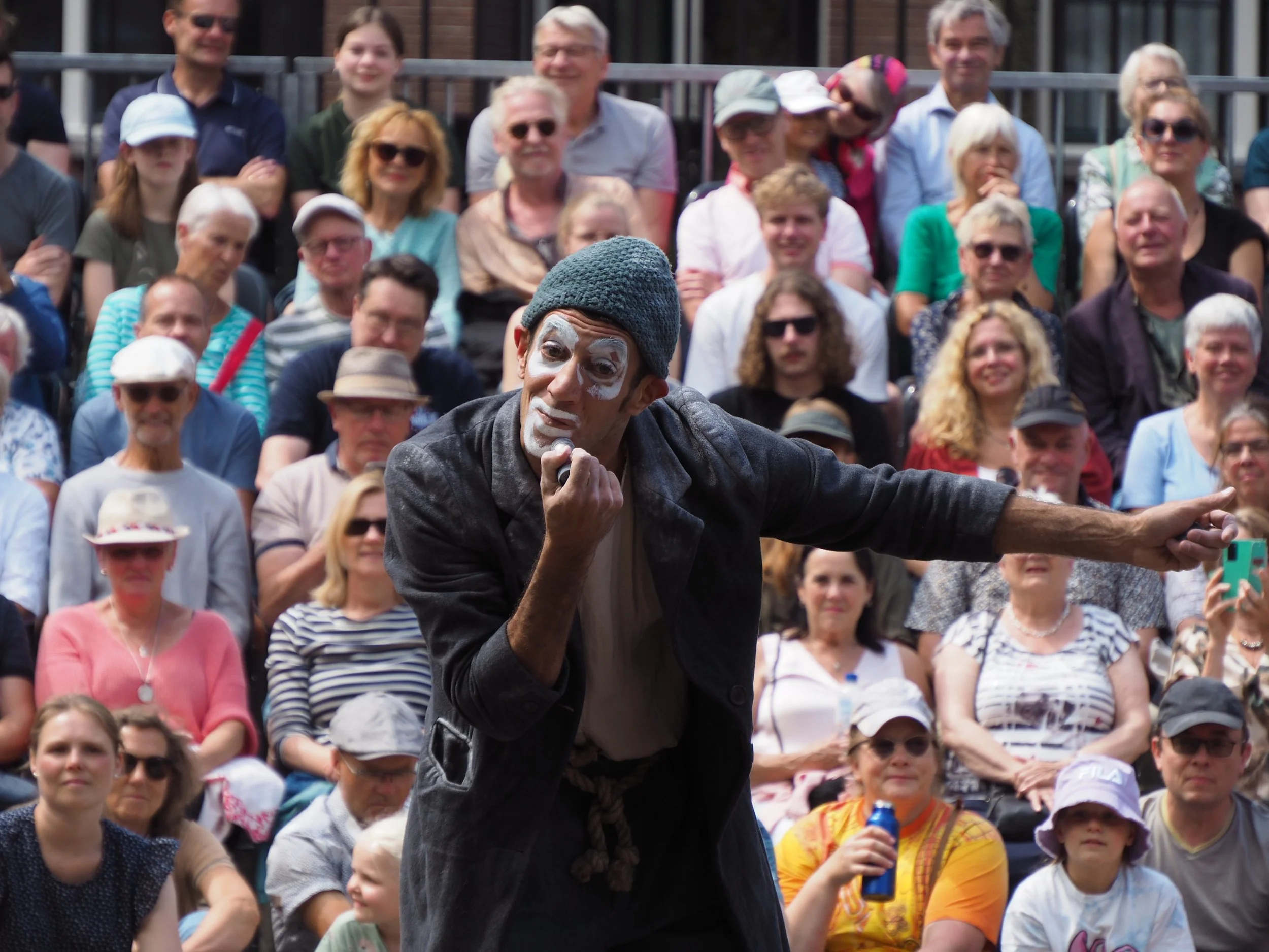 A street performer dressed as a mime with white face paint, wearing a gray hat and dark clothing, performing in front of a large audience sitting outdoors.