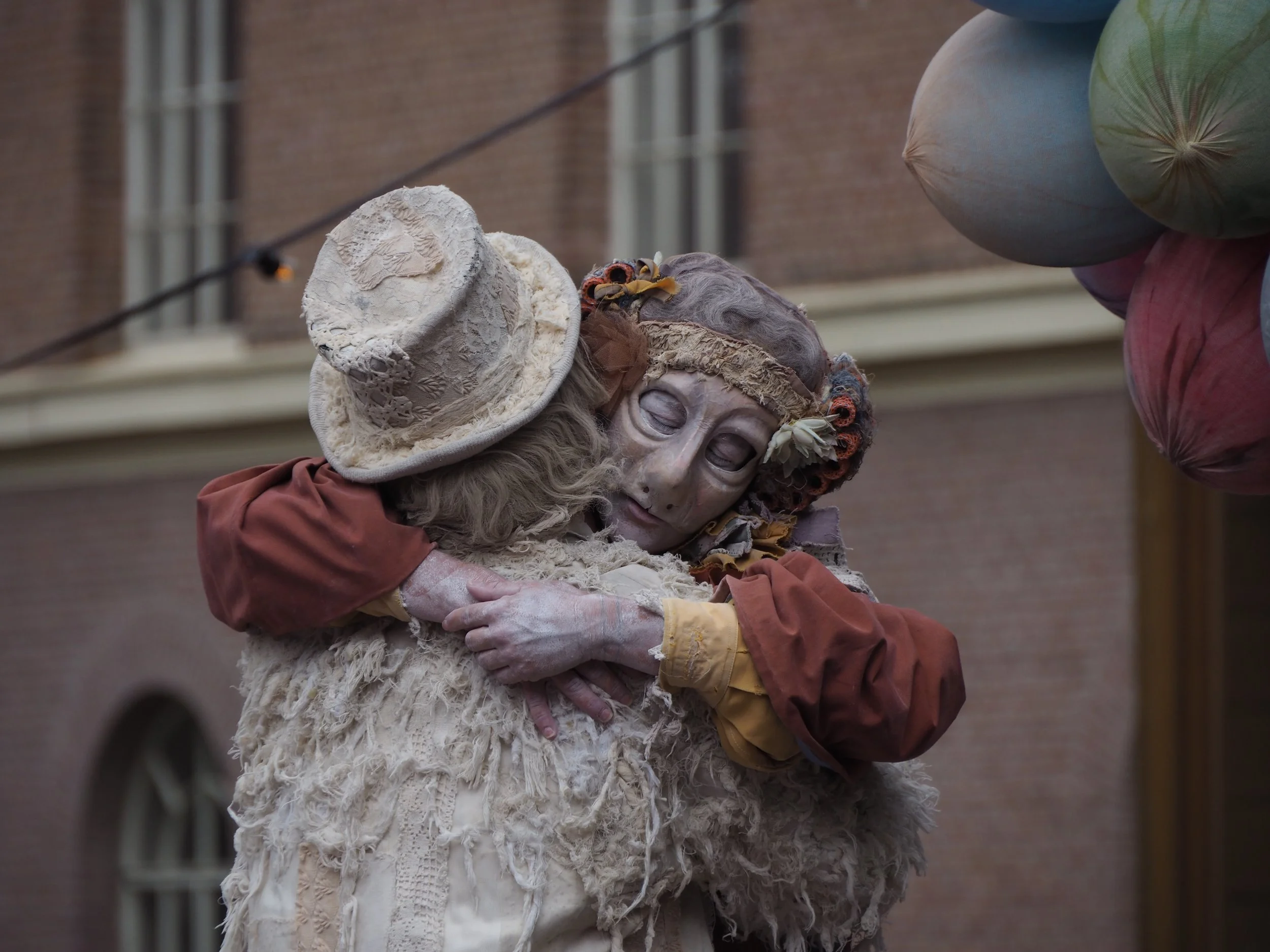 Two people dressed as old-fashioned characters embracing, one wearing a cream lace hat and the other wearing a headscarf and a mask with a painted face, in front of a brick building.