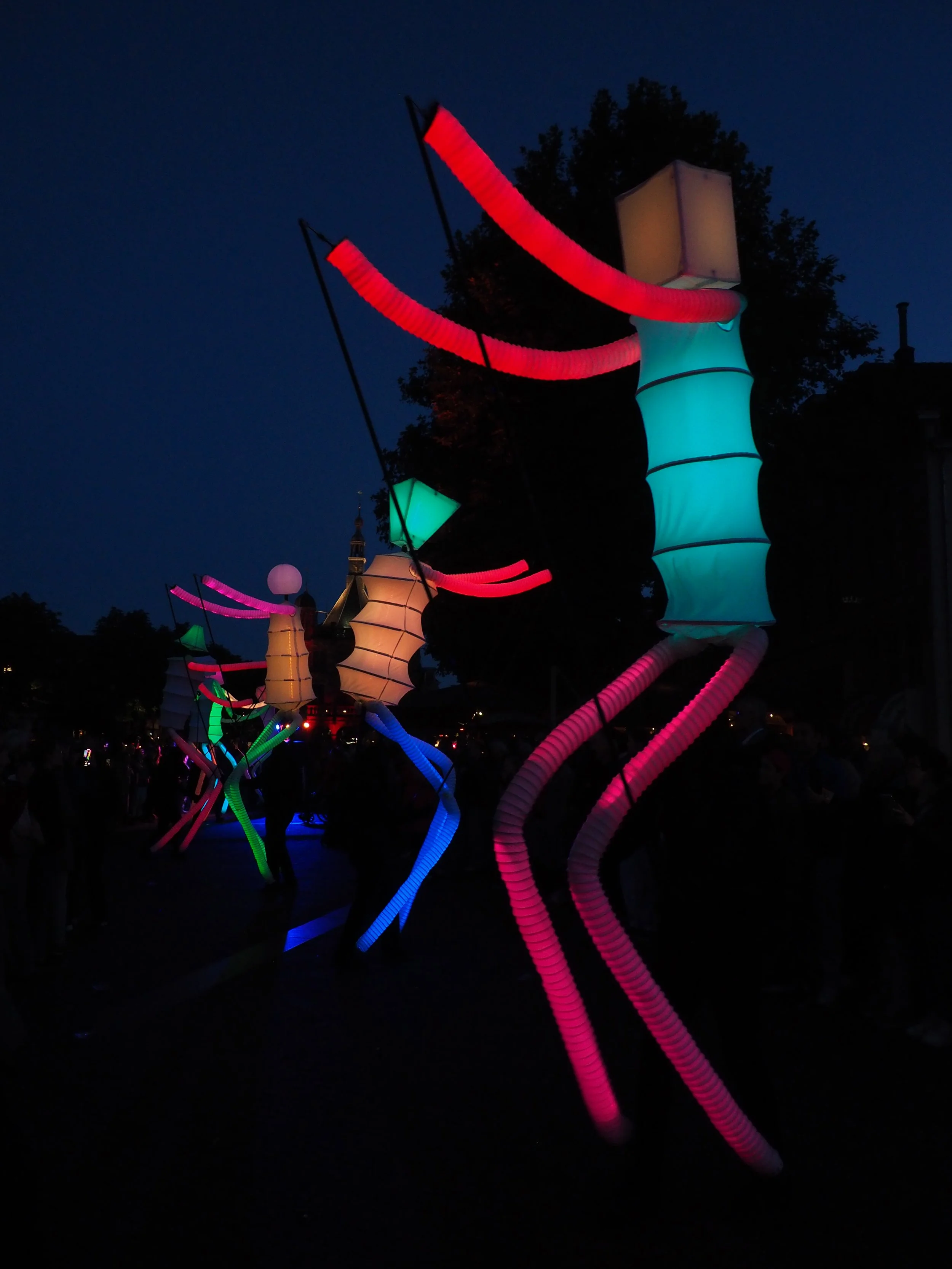 Colorful glowing lanterns and tubing illuminated by neon lights hanging over a crowd at night.