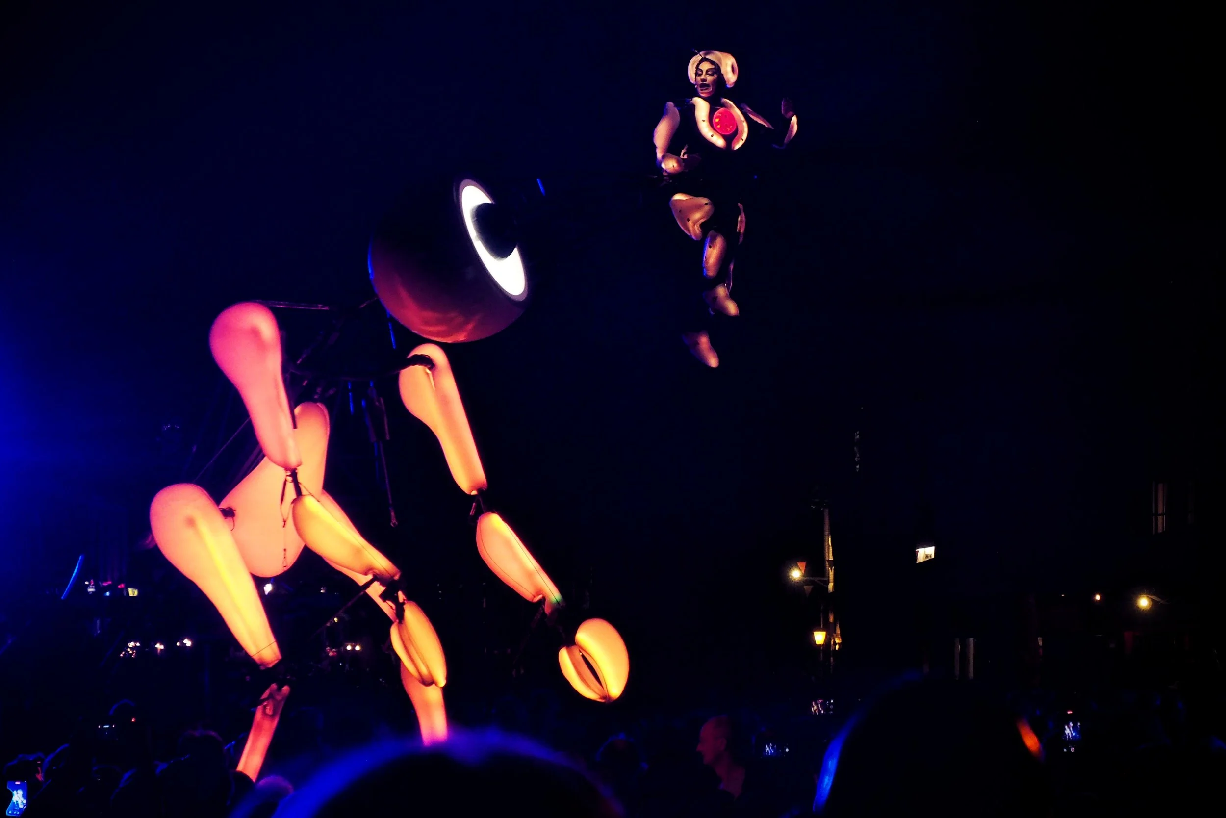 Nighttime scene featuring illuminated large sculptures of animals, including a dog and a kangaroo, with a person dressed as a clown or performer floating or jumping in the air.