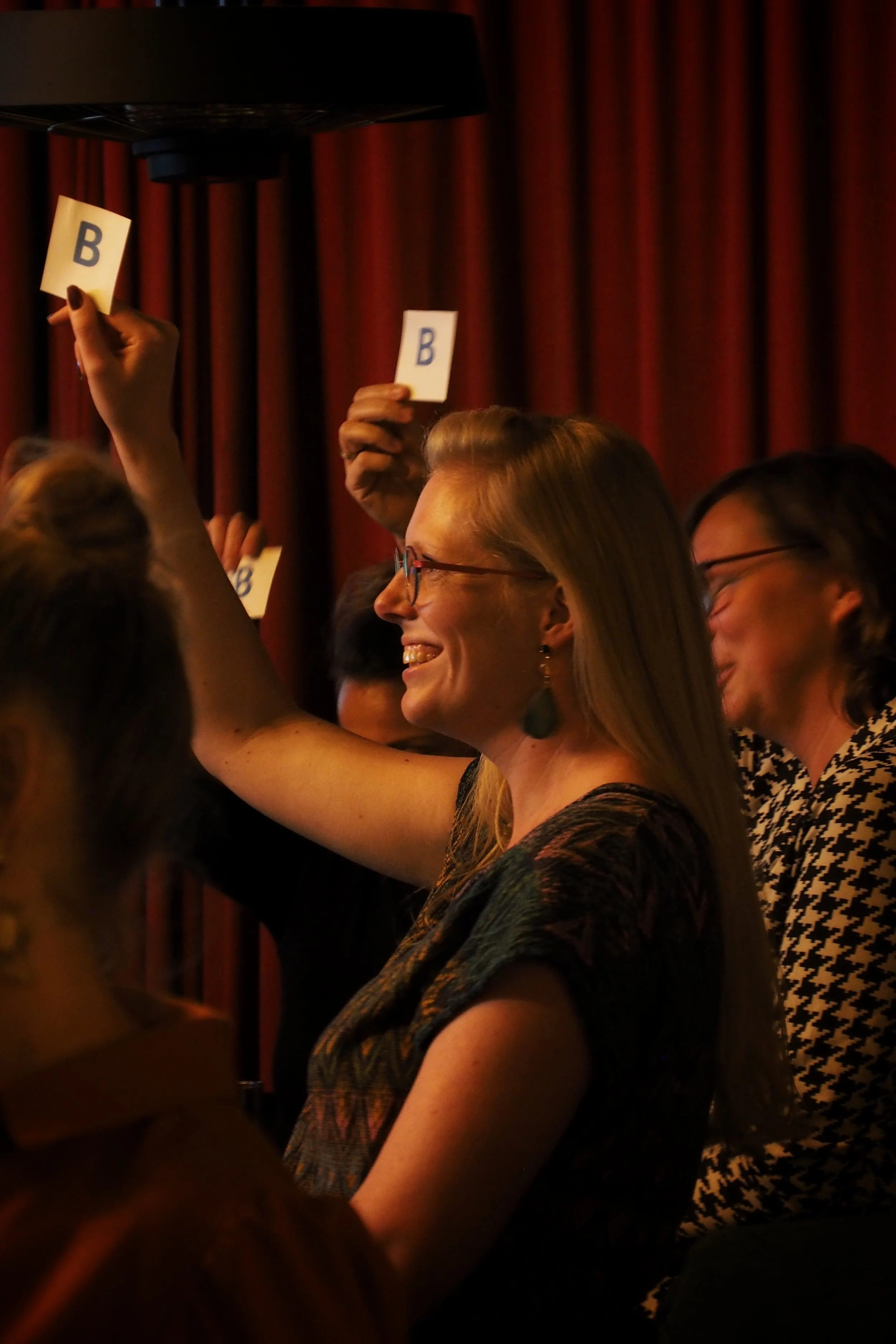 People in a room holding up small cards with the letter B on them, engaging in a group activity or voting, with a red curtain in the background.