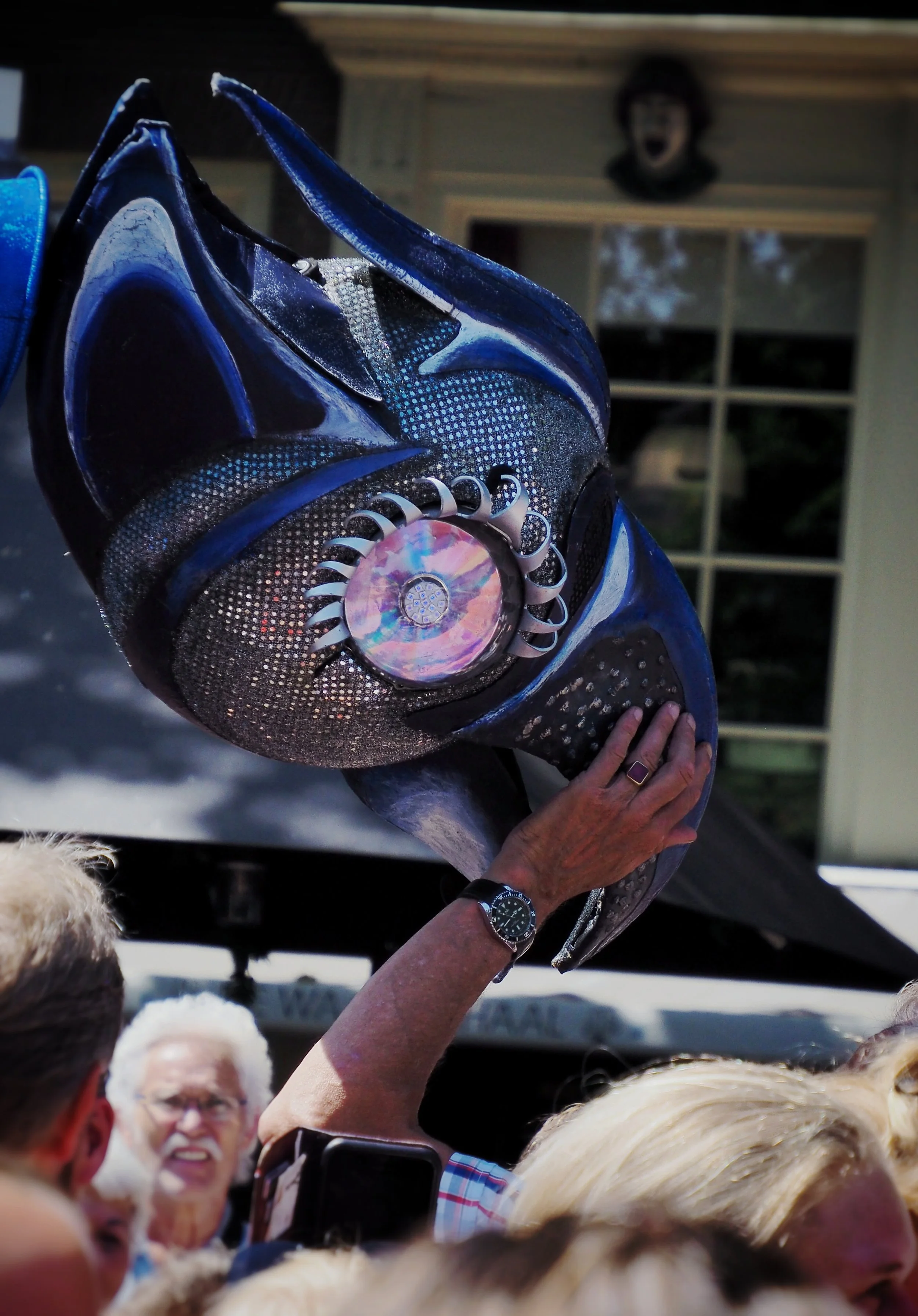 A person is holding a decorative black, blue, and silver mask with a large, iridescent jewel in the center, among a crowd of people with mostly blonde hair. In the background, a window and a person with a smiling face are visible.