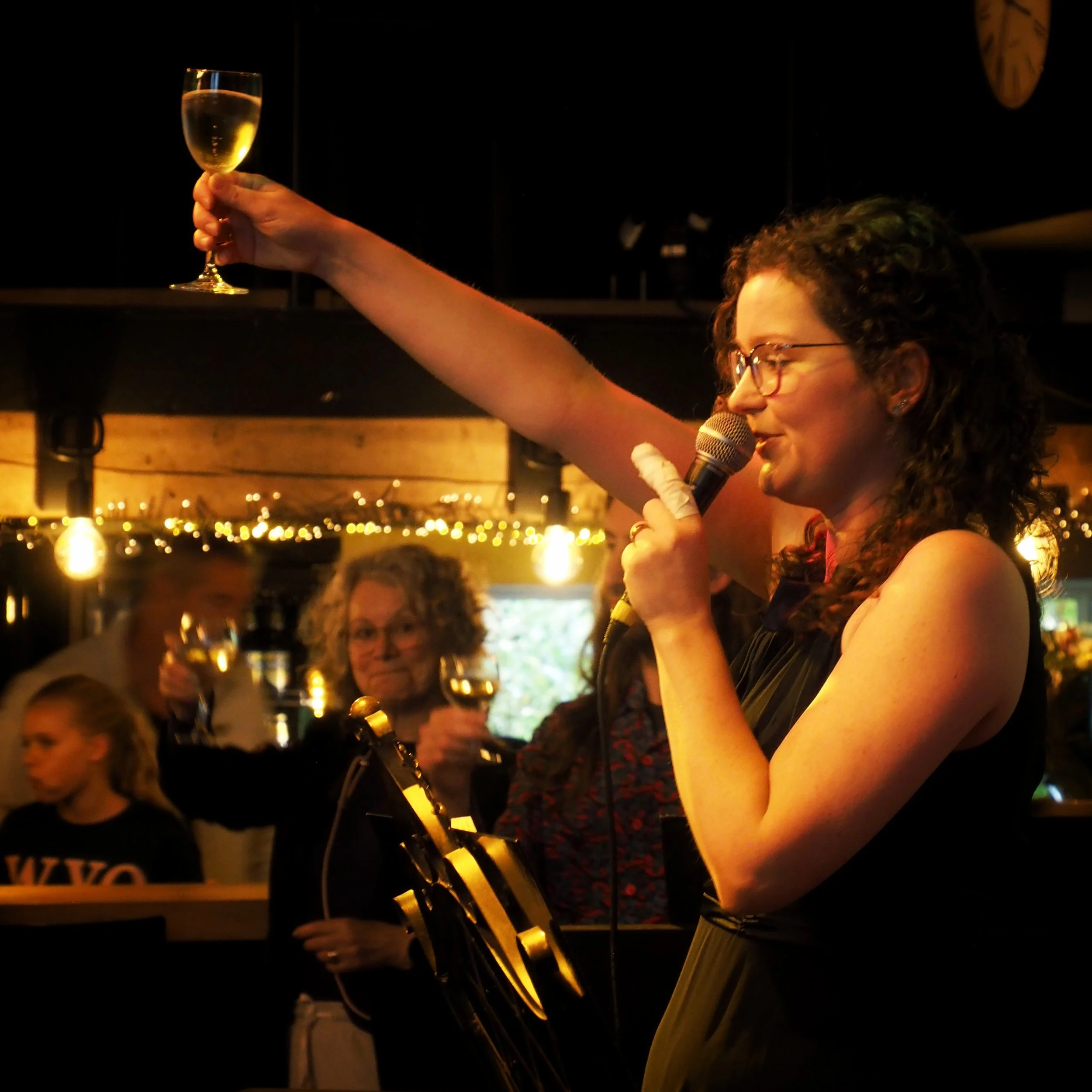 A person with curly hair and glasses holding a champagne flute at a celebration or party in a dimly lit venue with warm lighting and string lights.