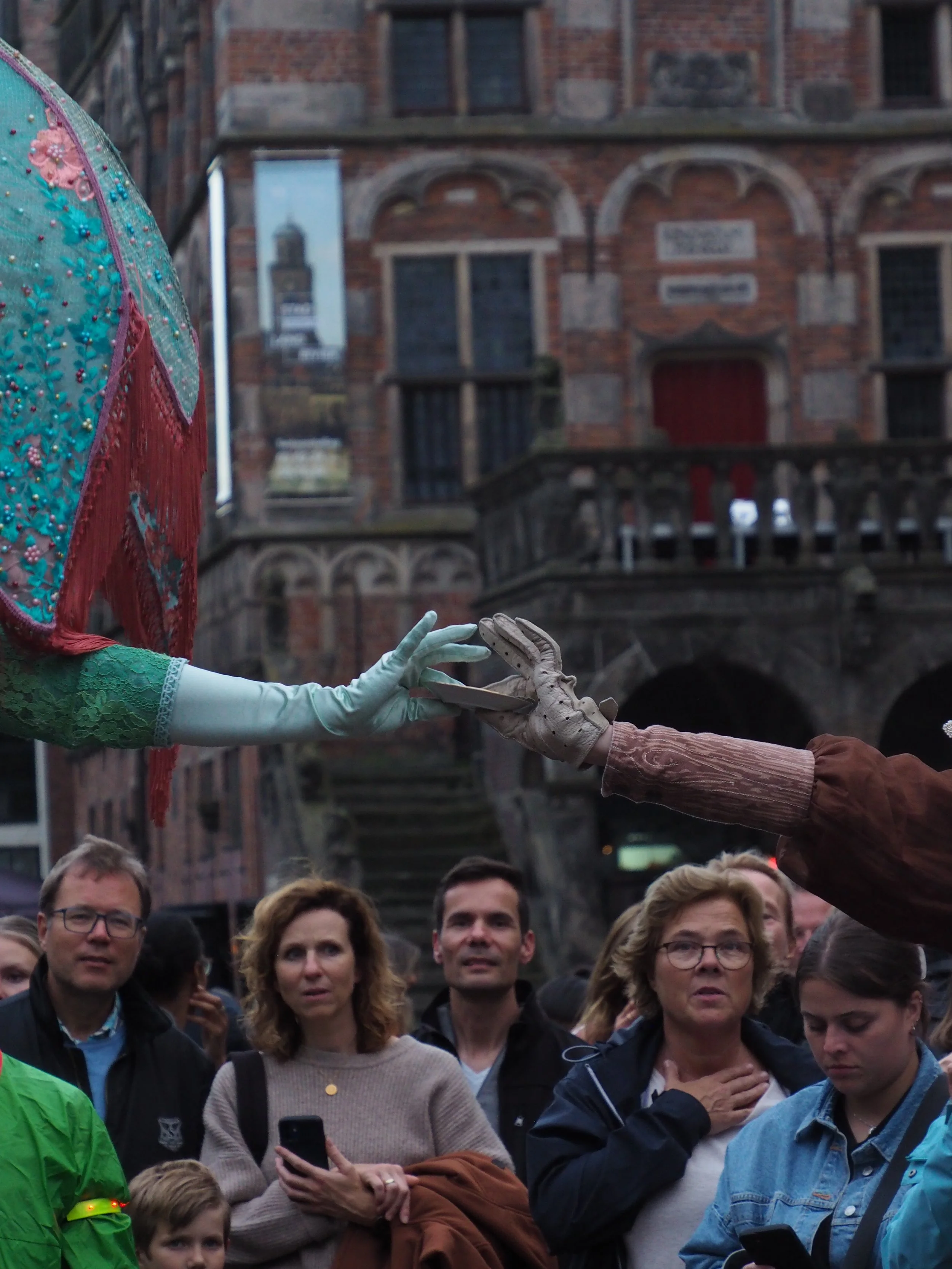 A group of people standing outdoors near a brick building, participating in a memorial or tribute event, with some individuals holding phones and placing their hands over their hearts.