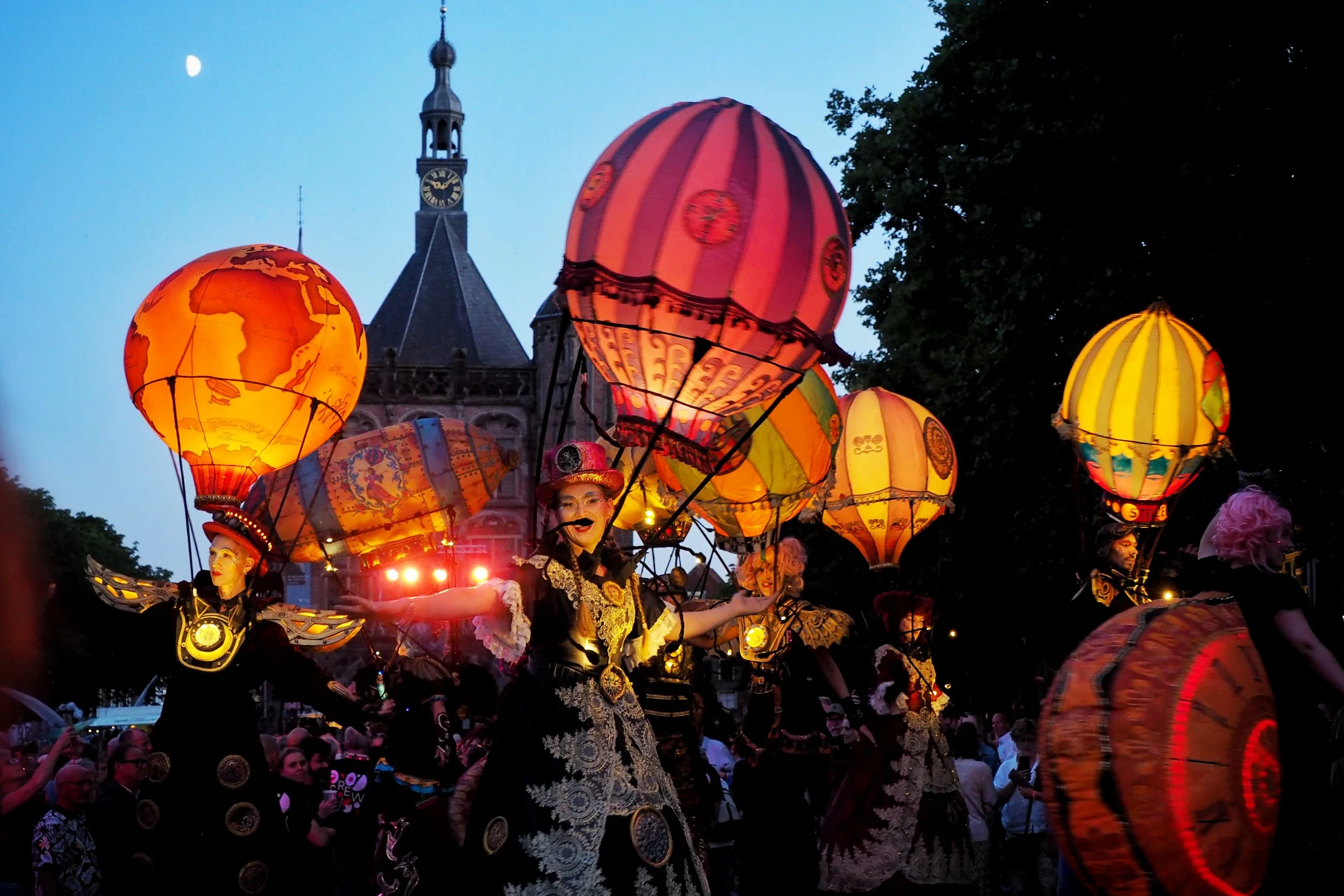 Nighttime parade with performers dressed in black and gold, holding illuminated lanterns shaped like globes and hot air balloons, with a historic clock tower and large crowd in the background.