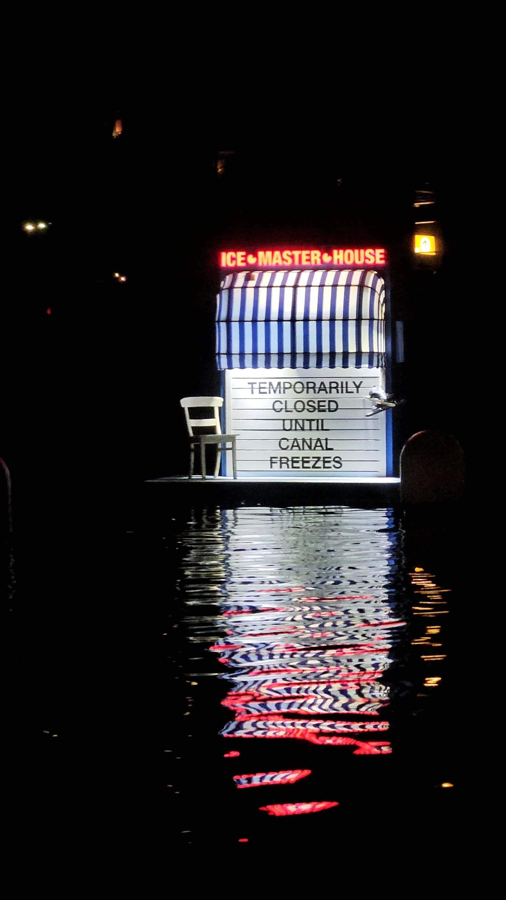 A signboard on a boat or dock indicating that the ice master house is temporarily closed until the canal freezes, with a chair next to the sign and a reflection of the sign in the water.