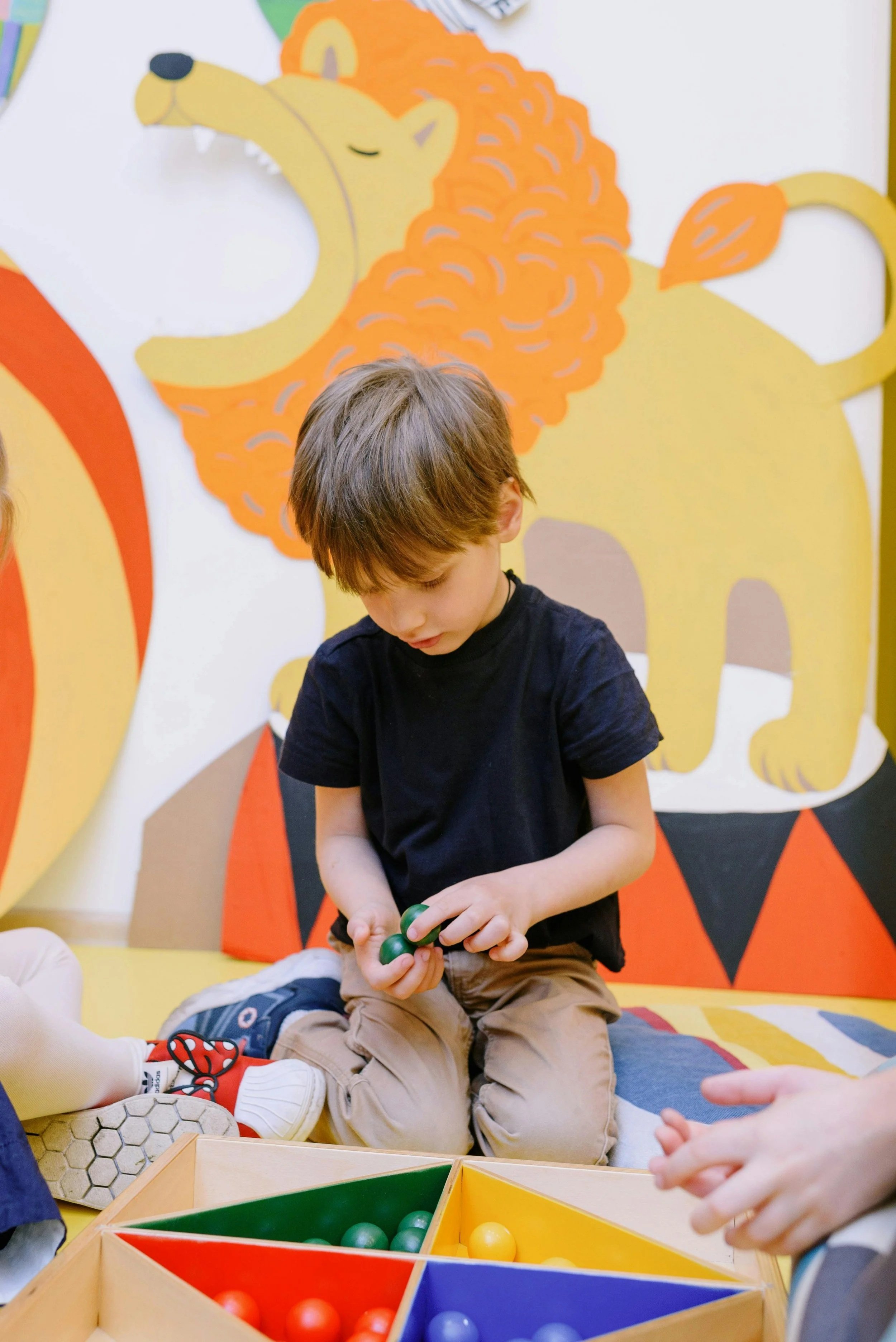 A young boy sitting on a colorful mat, holding green balls, in front of a painted lion mural with an orange mane.