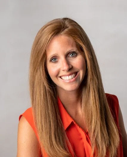 A woman with long red hair smiling, wearing a sleeveless orange top, against a plain light gray background.