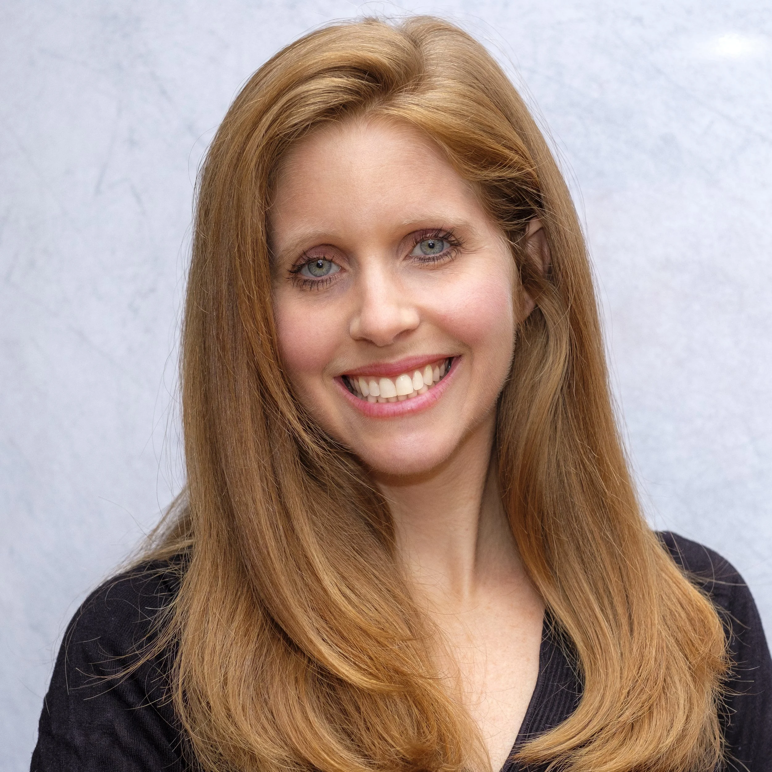 A portrait of a smiling woman with long red hair, blue eyes, and fair skin, wearing a black top, against a neutral background.