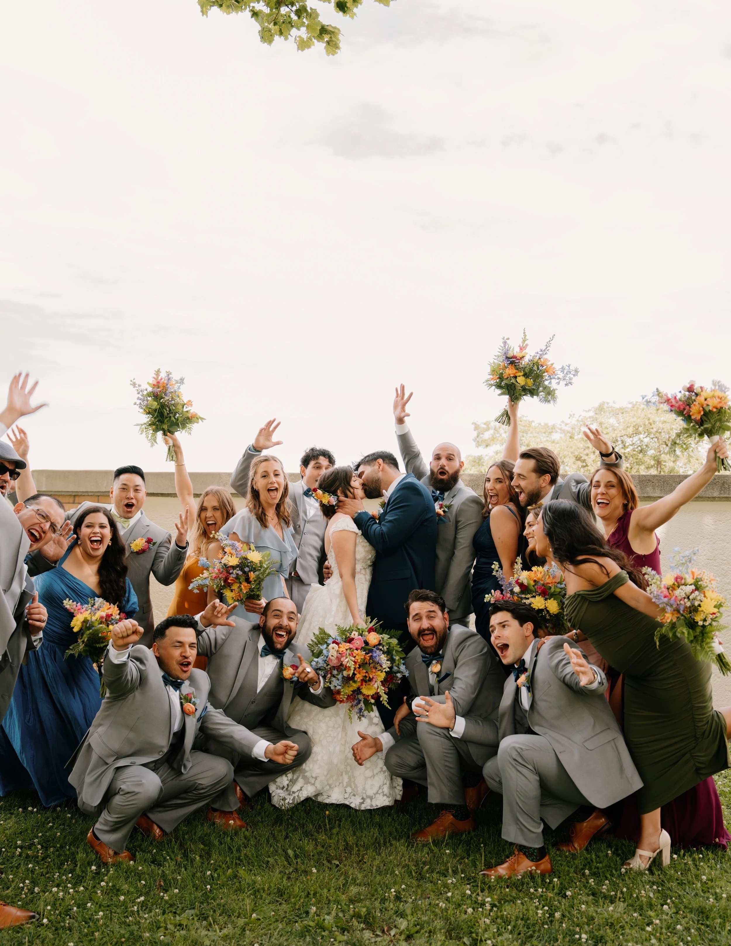 A large group of wedding guests celebrating outdoors, with a newlywed couple in the center sharing a kiss, surrounded by friends holding bouquets, raising hands, and smiling.