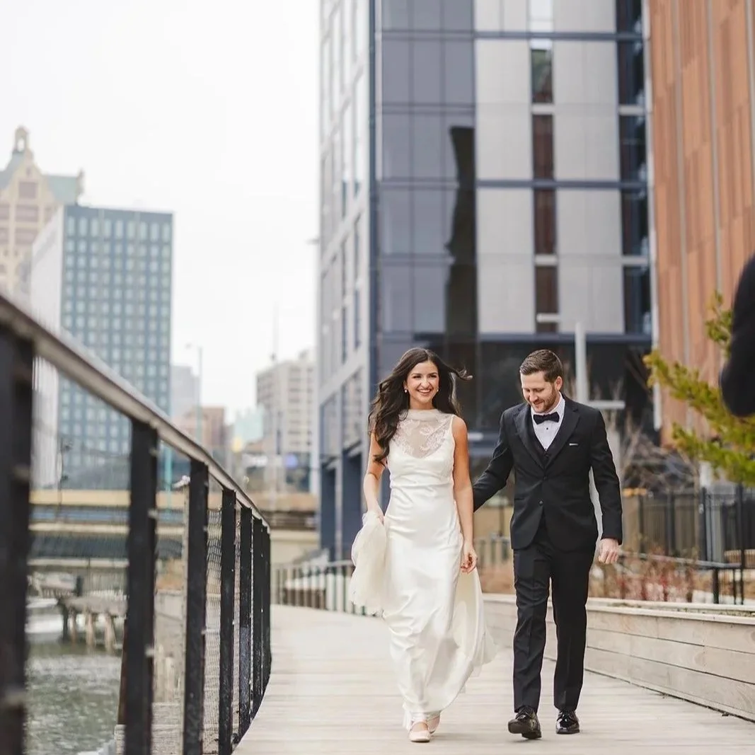 A happy couple in wedding attire walking hand in hand on a city sidewalk near a waterway, with tall modern buildings in the background.