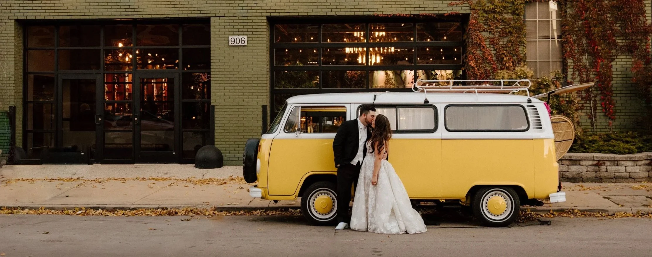 A couple dressed in wedding attire sharing a kiss in front of a yellow vintage van parked on a city street, with a green brick building and autumn leaves in the background.