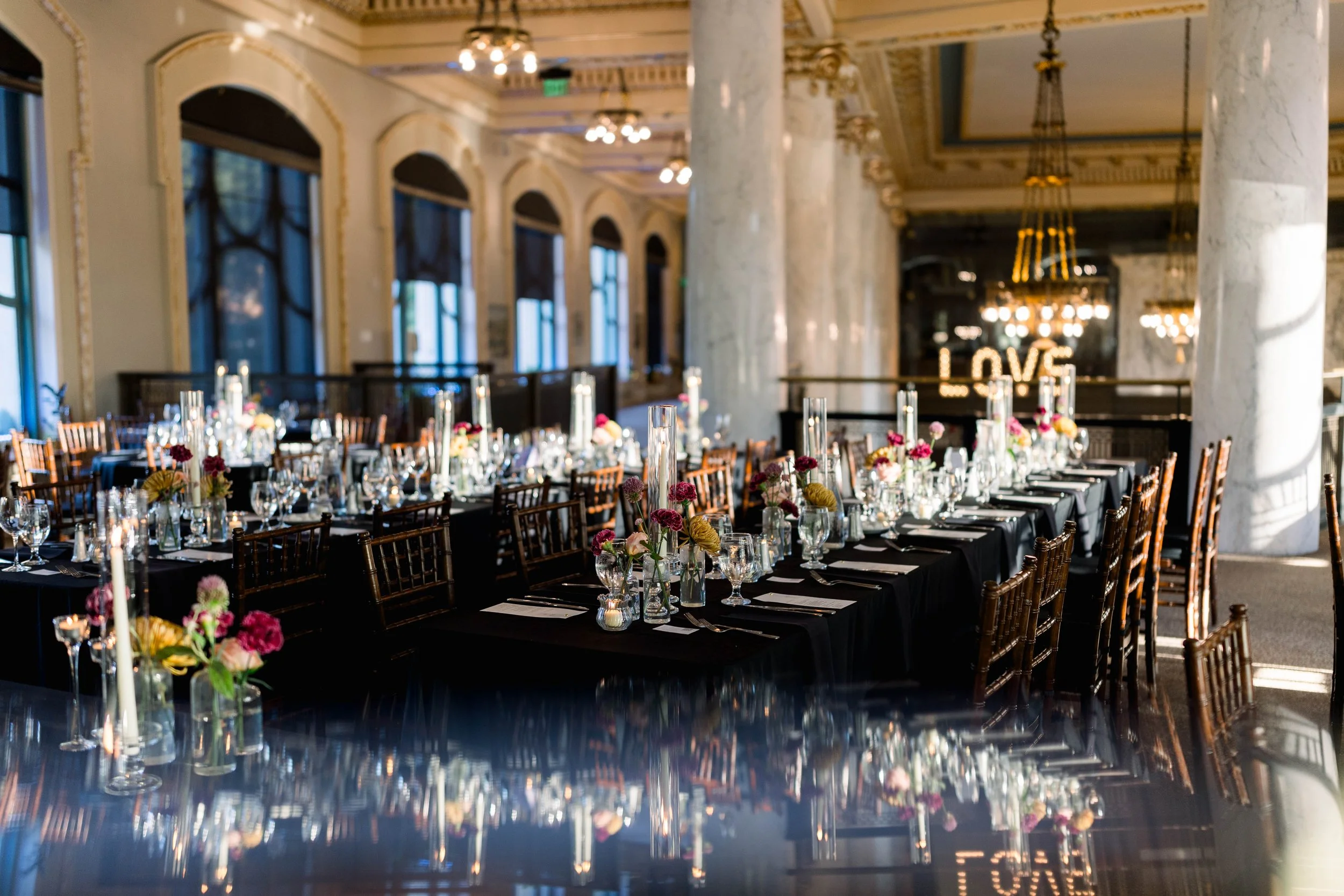 Elegant banquet hall with large windows, marble columns, and ornate ceiling details. Decorated tables with black tablecloths, plates, glassware, candles, and pink and yellow floral centerpieces. Illuminated 'LOVE' sign in the background.