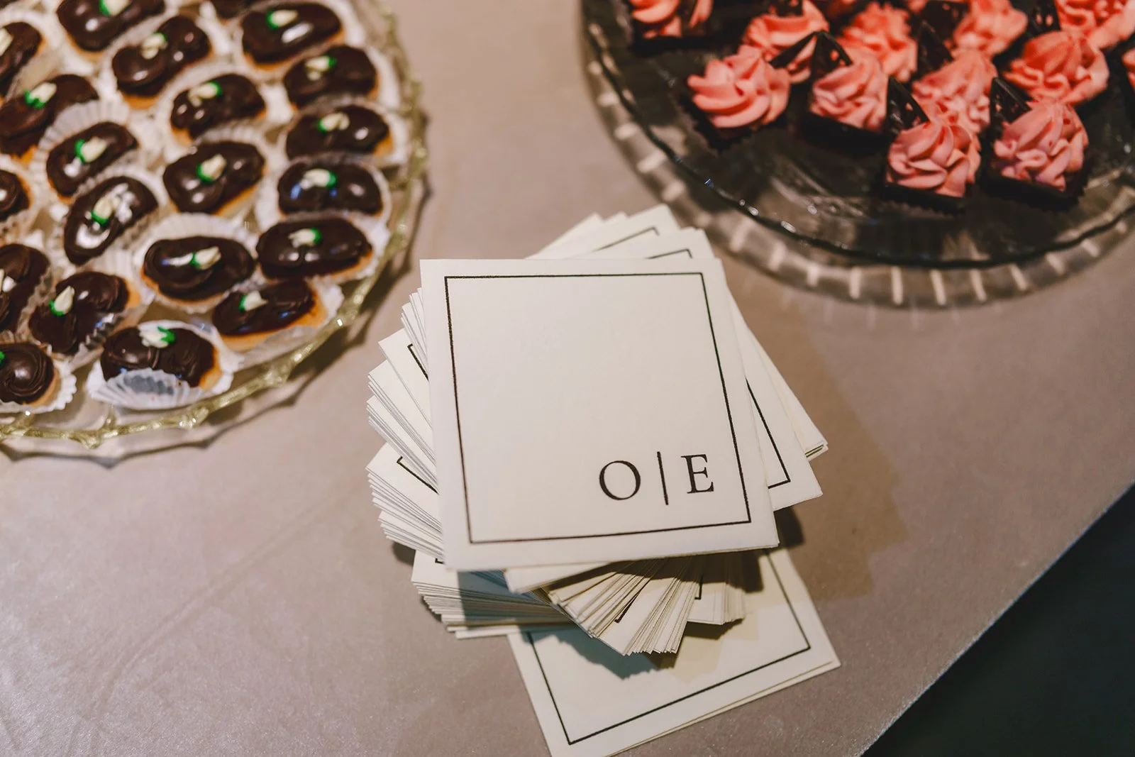 A table with plates of decorated mini cupcakes and pink cupcakes, with a stack of blank cards labeled 'LOVE' in black text.