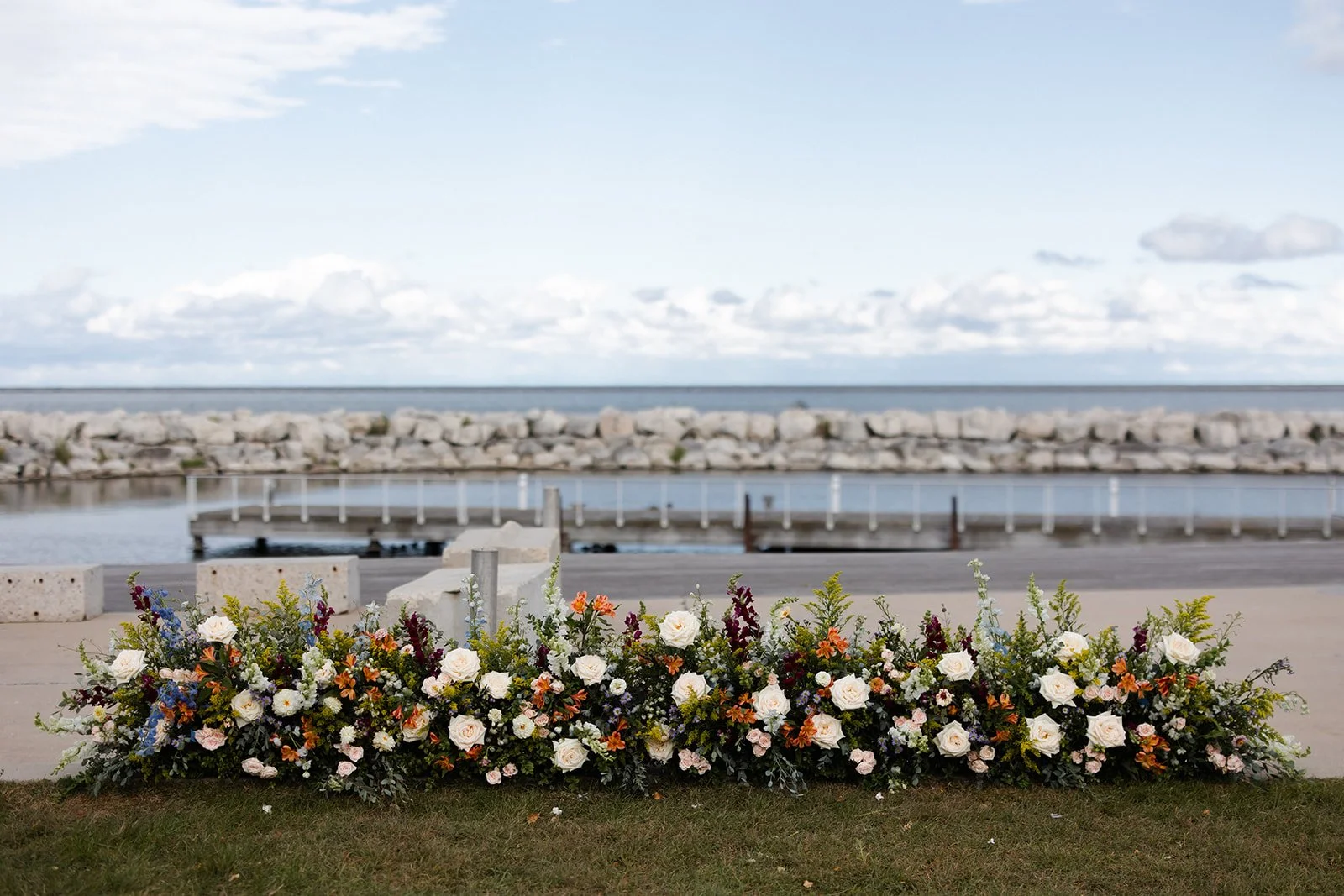 A floral arrangement with white, orange, purple, and pink flowers in front of a body of water and a rocky shoreline under a partly cloudy sky.