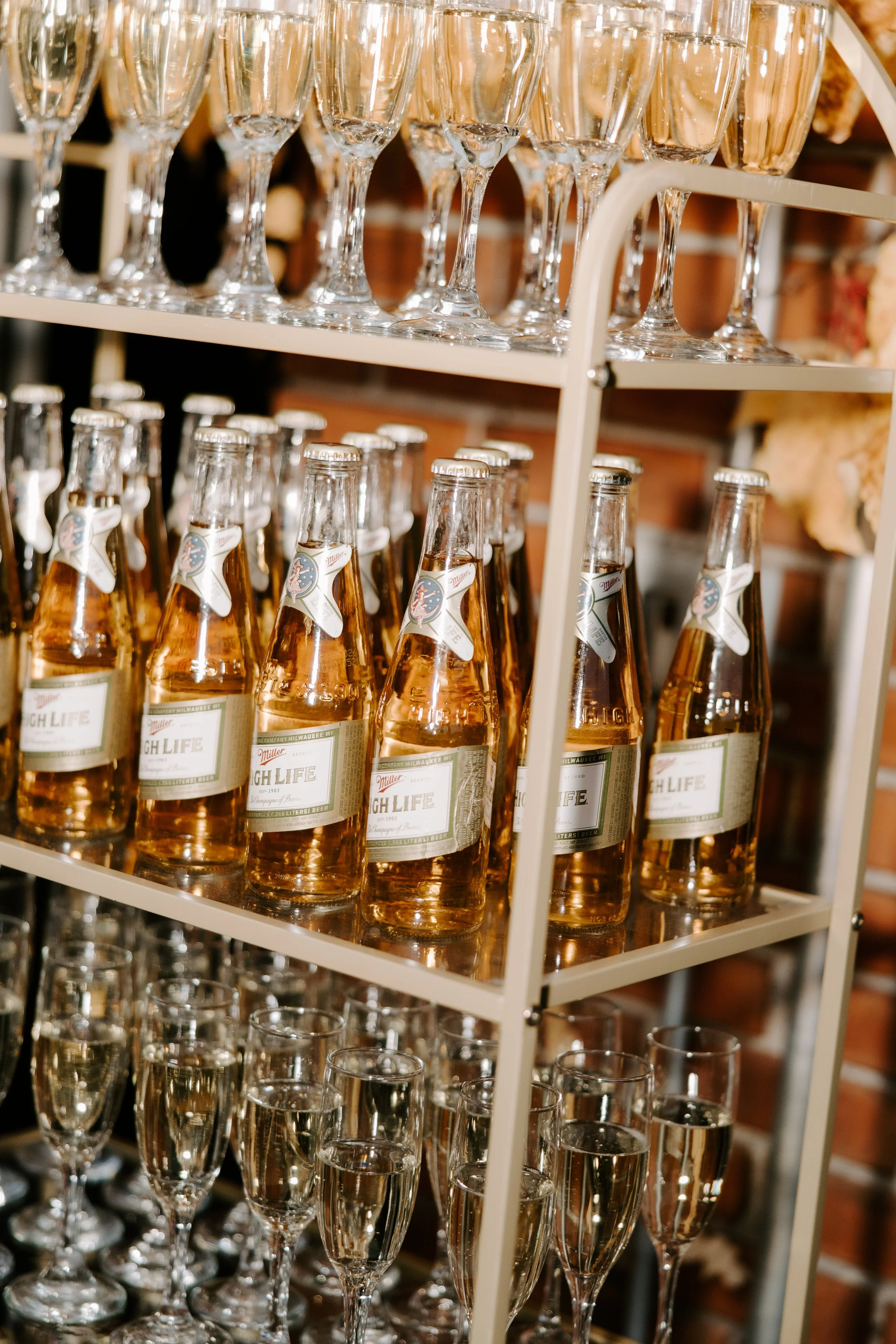 A white shelving unit holding rows of champagne glasses on top and Miller High Life beer bottles below.