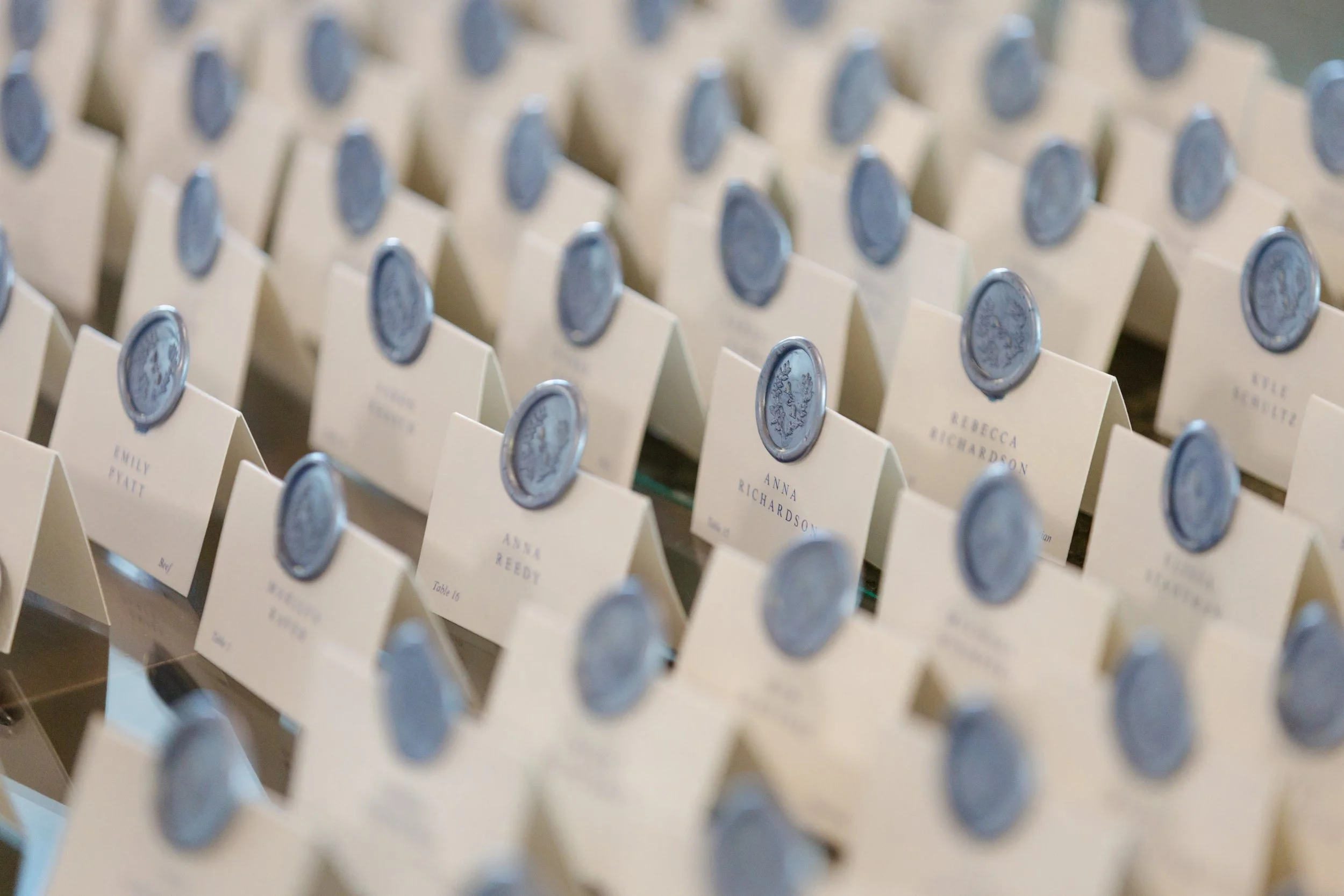 Wedding reception seating chart with small place cards on a whiteboard. Each card has a blue wax seal sticker and a guest's name printed underneath, such as Emily Pratt, Anna Richardson, and Rebecc Reedy.