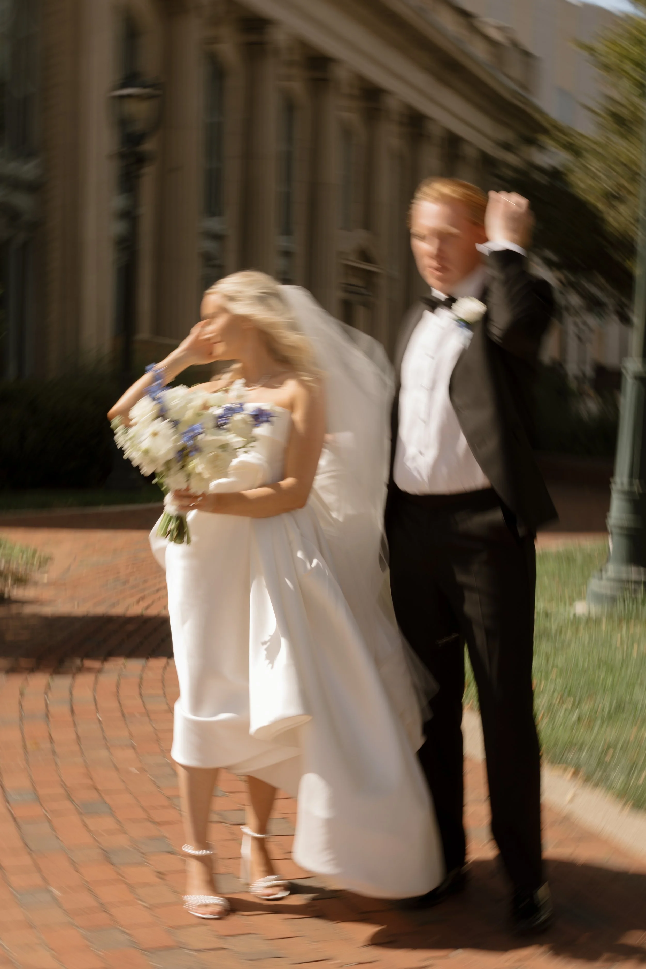 A bride and groom walking outside on a brick sidewalk in front of a building with tall windows. The groom is wearing a tuxedo and the bride is in a white wedding dress holding a bouquet of white flowers. The image is blurred.
