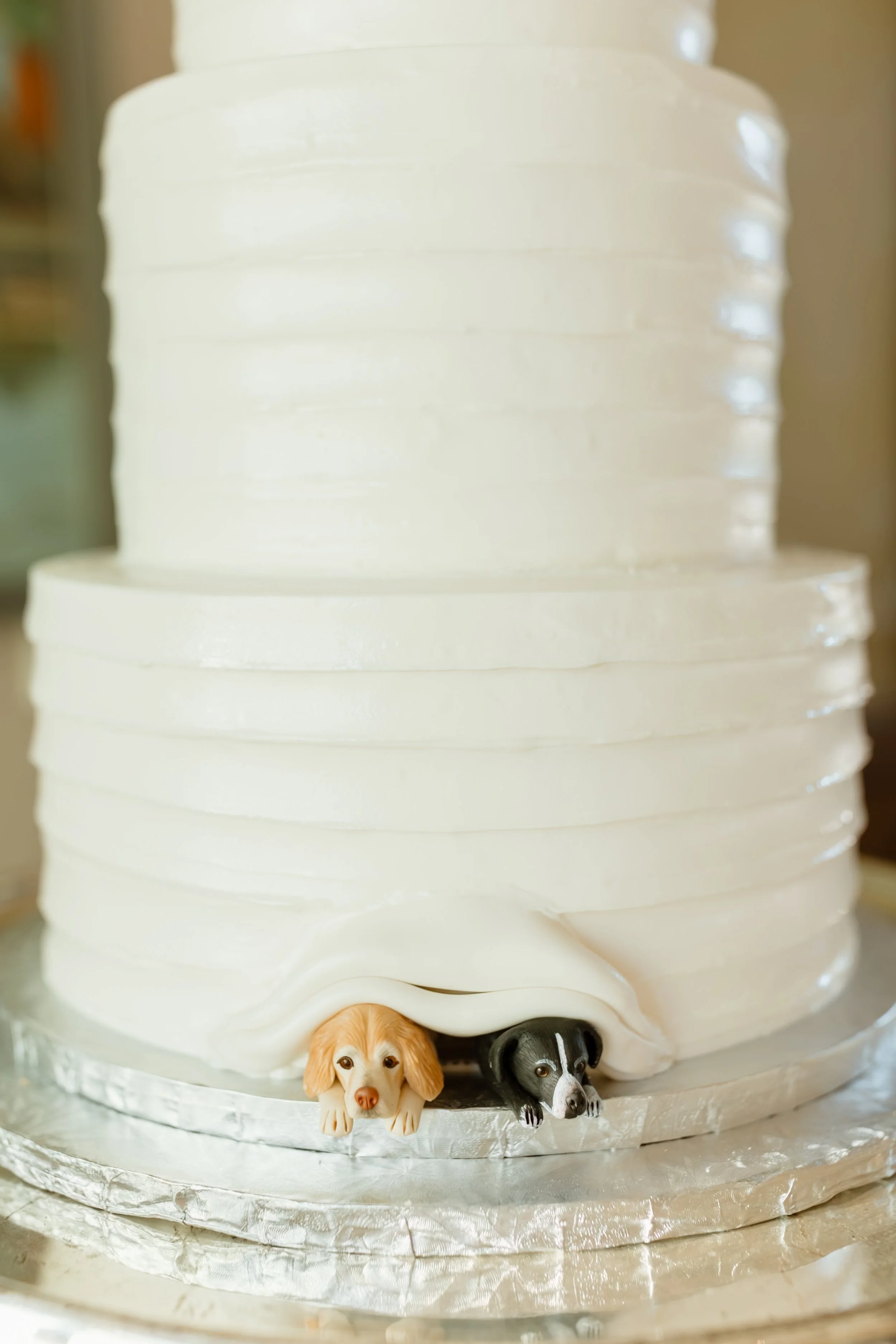 A white, multi-layered cake with dog figurines at the base, featuring a golden retriever, a black and white dog, and a black dog.