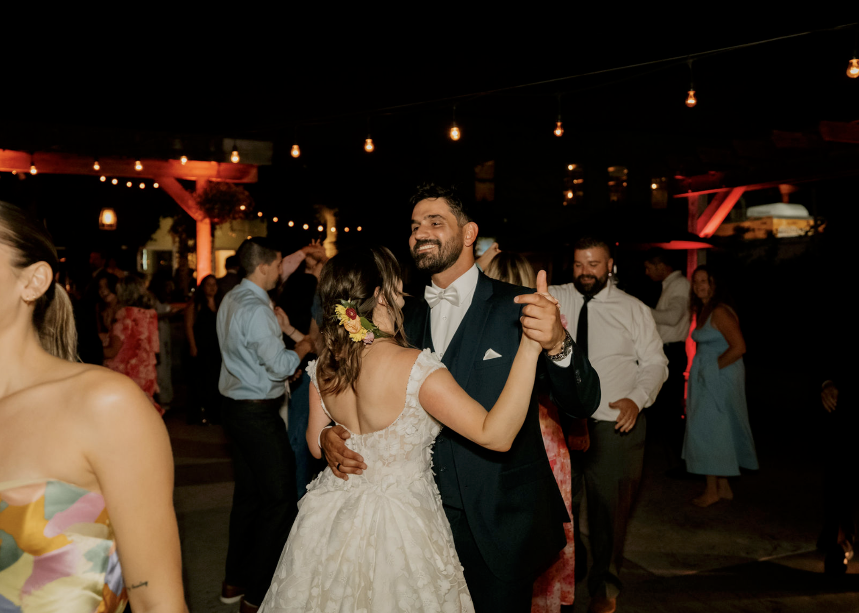 Couple dancing at wedding reception at night with string lights and guests in background.