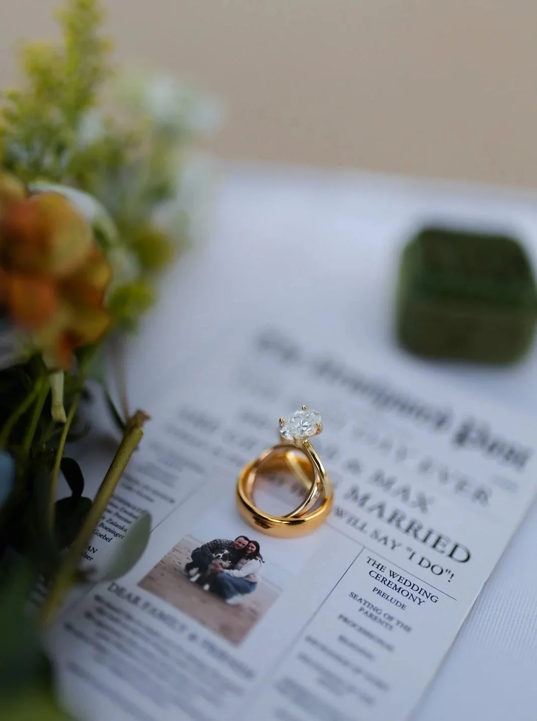 A wedding invitation with a gold engagement ring on top, accompanied by flowers and a blurred green object in the background.