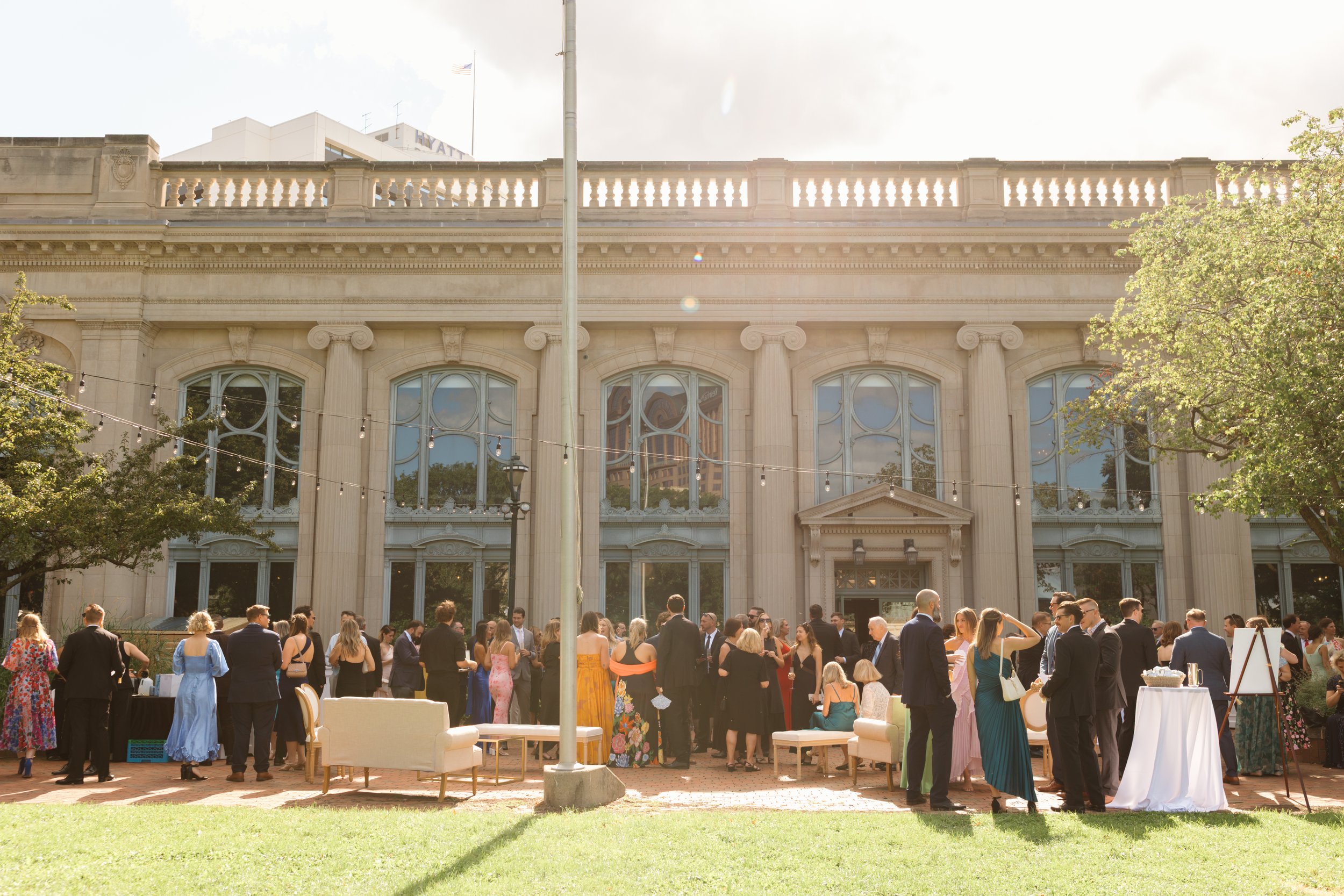 Wedding at The Society by Two Birds Event Group. People attending a formal outdoor event or wedding reception in front of a historic building with large windows, columns, and a stone facade, during daytime with sunny weather.