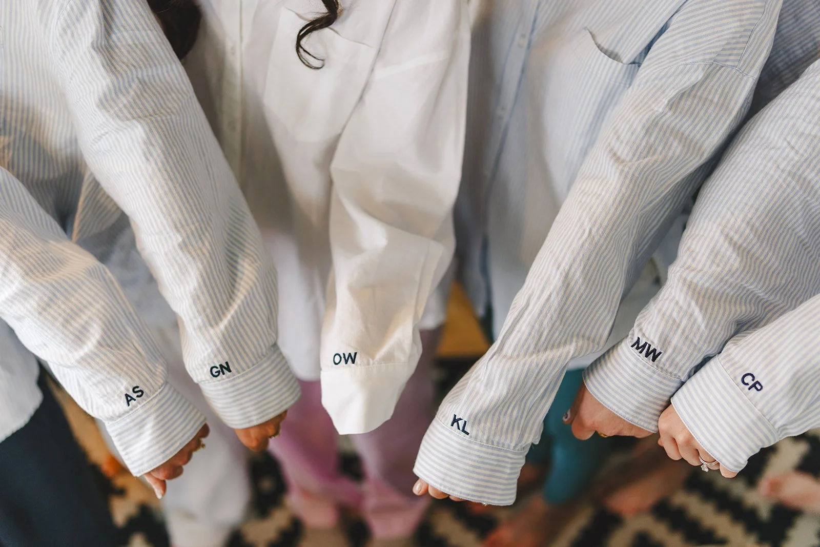Close-up of six people holding hands in a circle, all wearing white and blue striped shirts with initials embroidered on the sleeve cuffs.