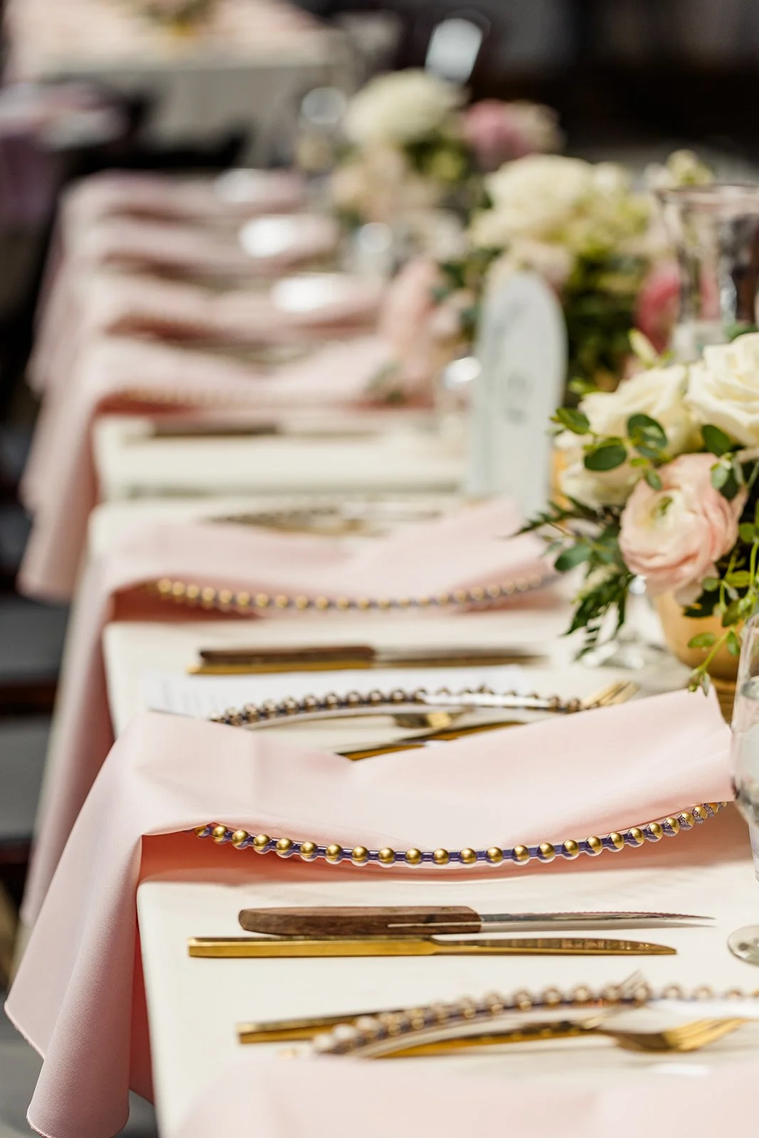 A long dining table set for a formal event with pink napkins, gold utensils, and floral centerpieces with white and pink roses.