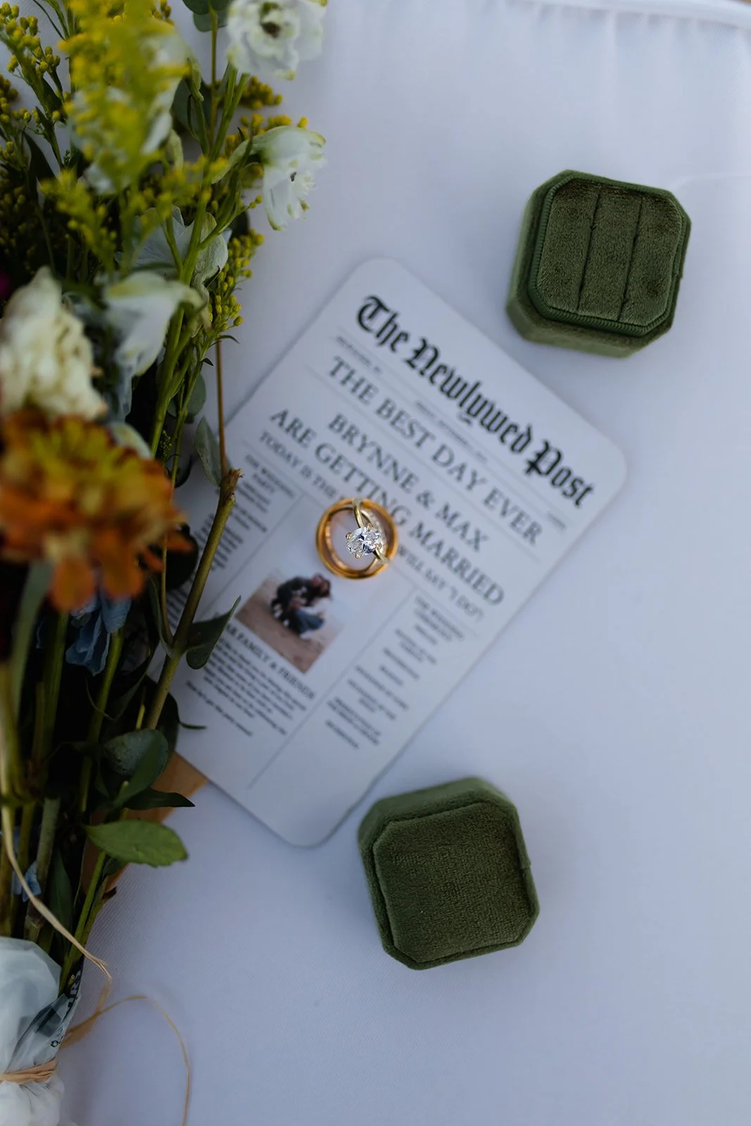 A wedding invitation, two green ring boxes, a bridal ring, and a bouquet of flowers on a white tablecloth.