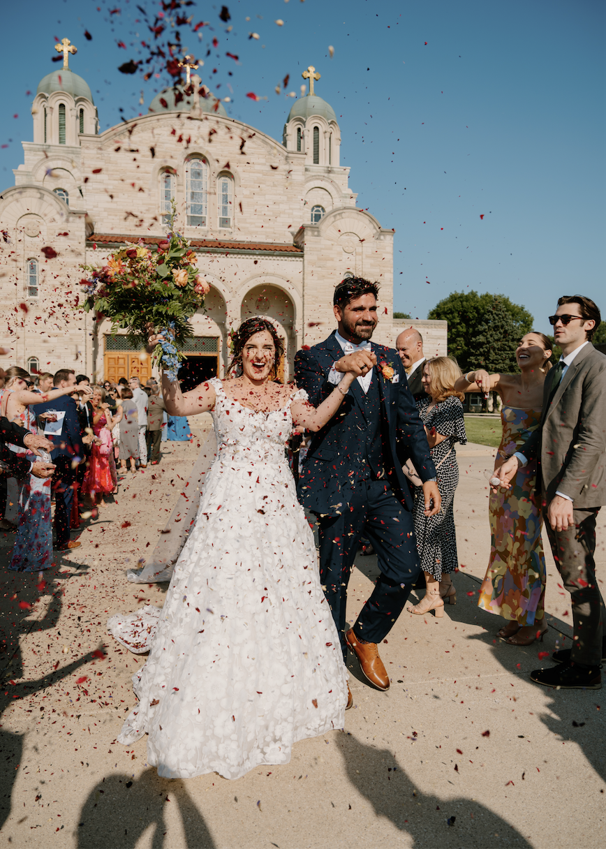 Happy newlywed couple walking hand in hand out of a church surrounded by friends and family, celebrating with confetti on a sunny day.