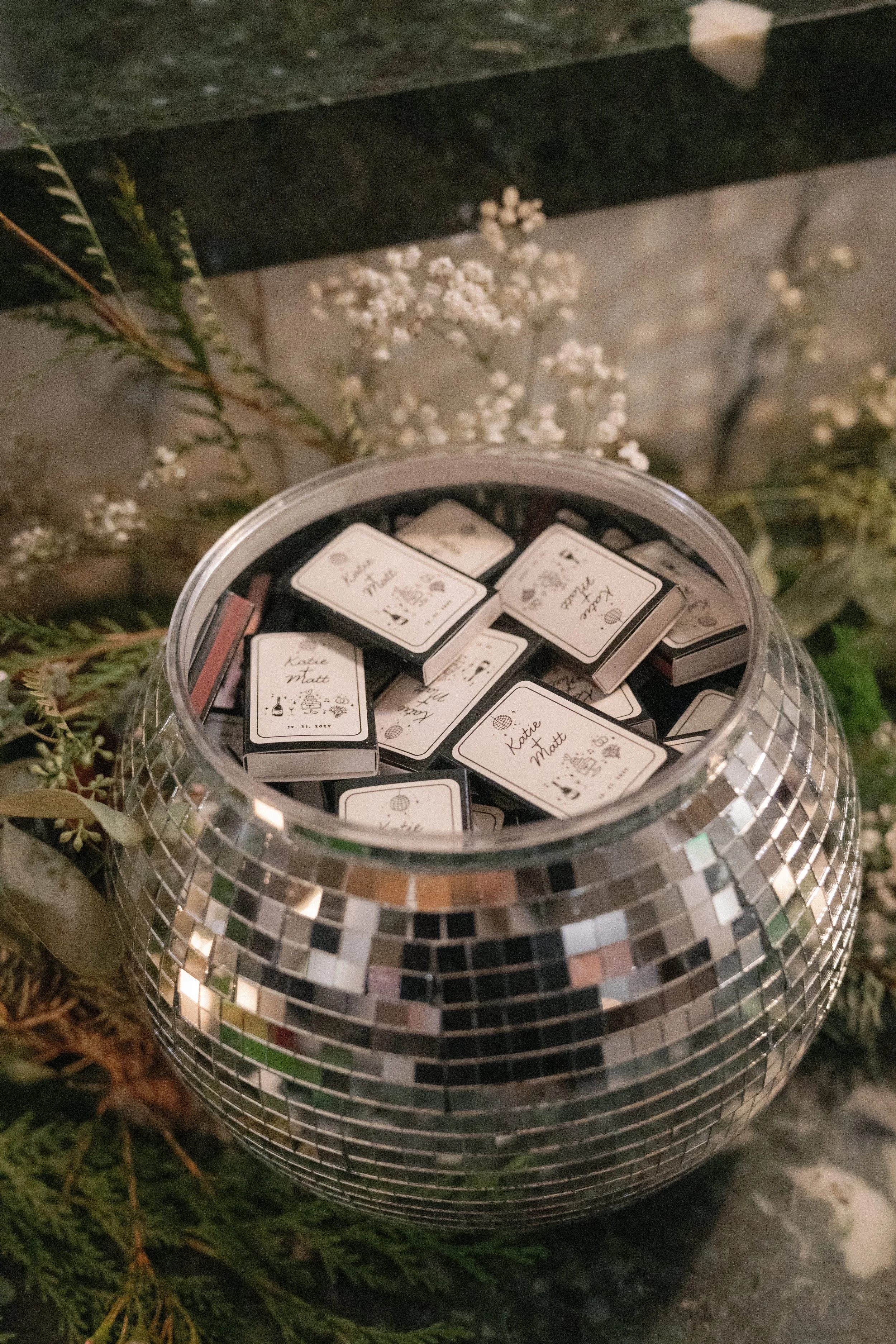 A clear plastic jar filled with small matchboxes, each labeled with a name, placed on a reflective metallic surface with a mosaic pattern. There are plants and a dark stone surface in the background.