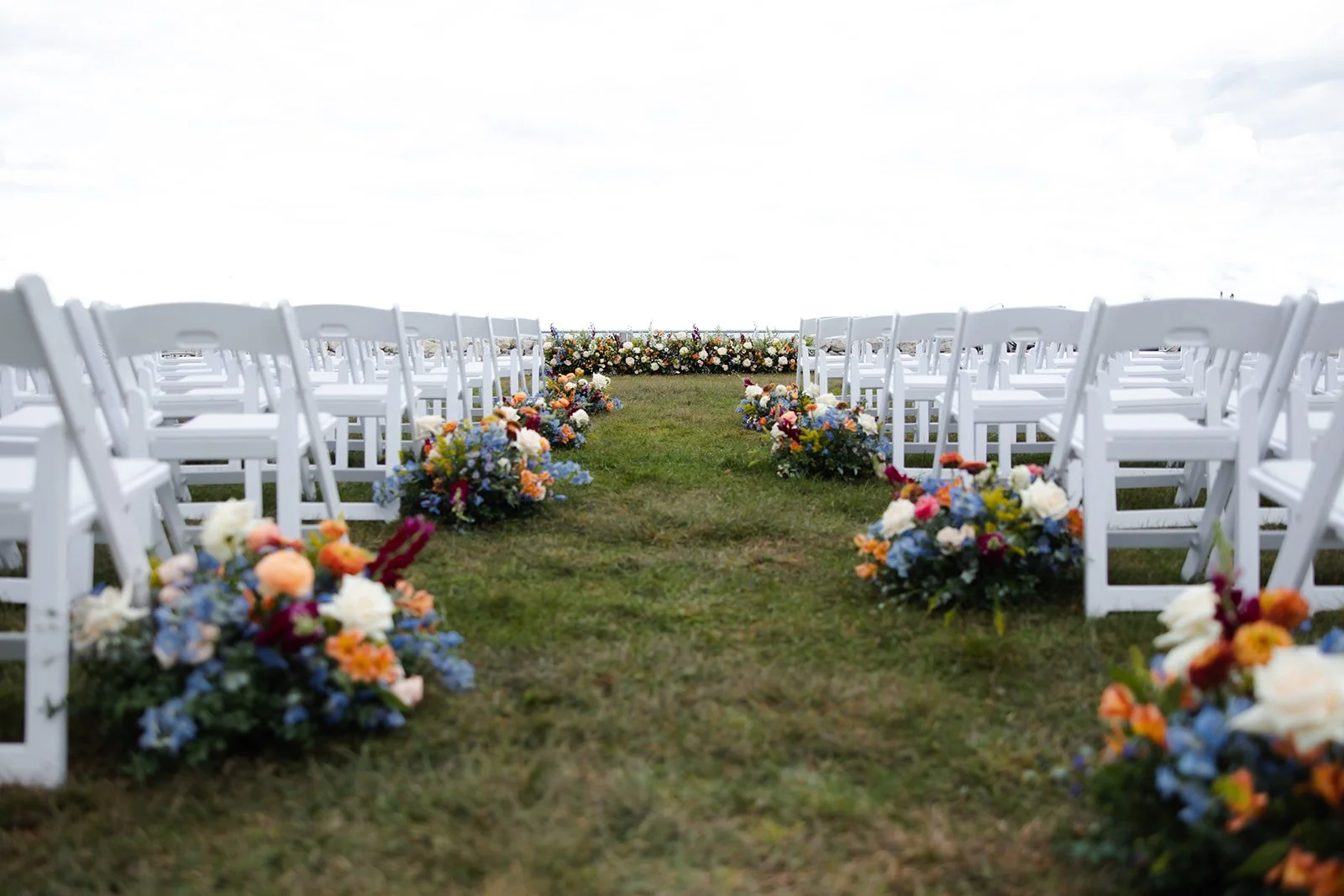 Wedding ceremony setup with white chairs and colorful floral arrangements along the aisle on a grassy outdoor field under a cloudy sky.