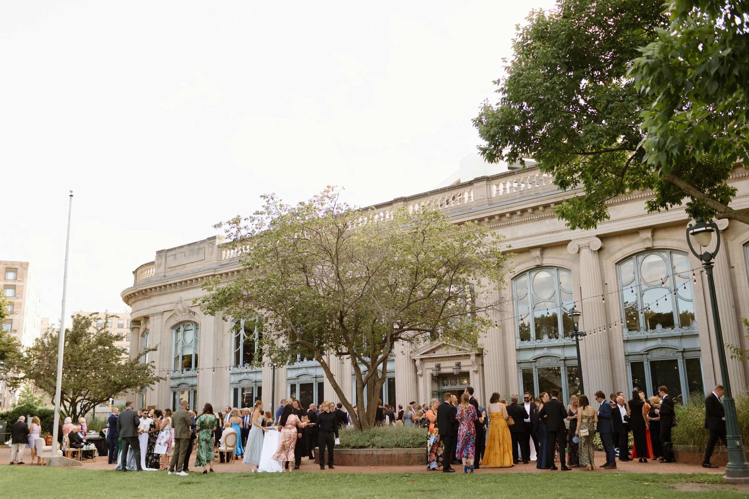 People in formal attire gathered outside a historic building, likely for a wedding or special event, with trees, string lights, and a cloudy sky.