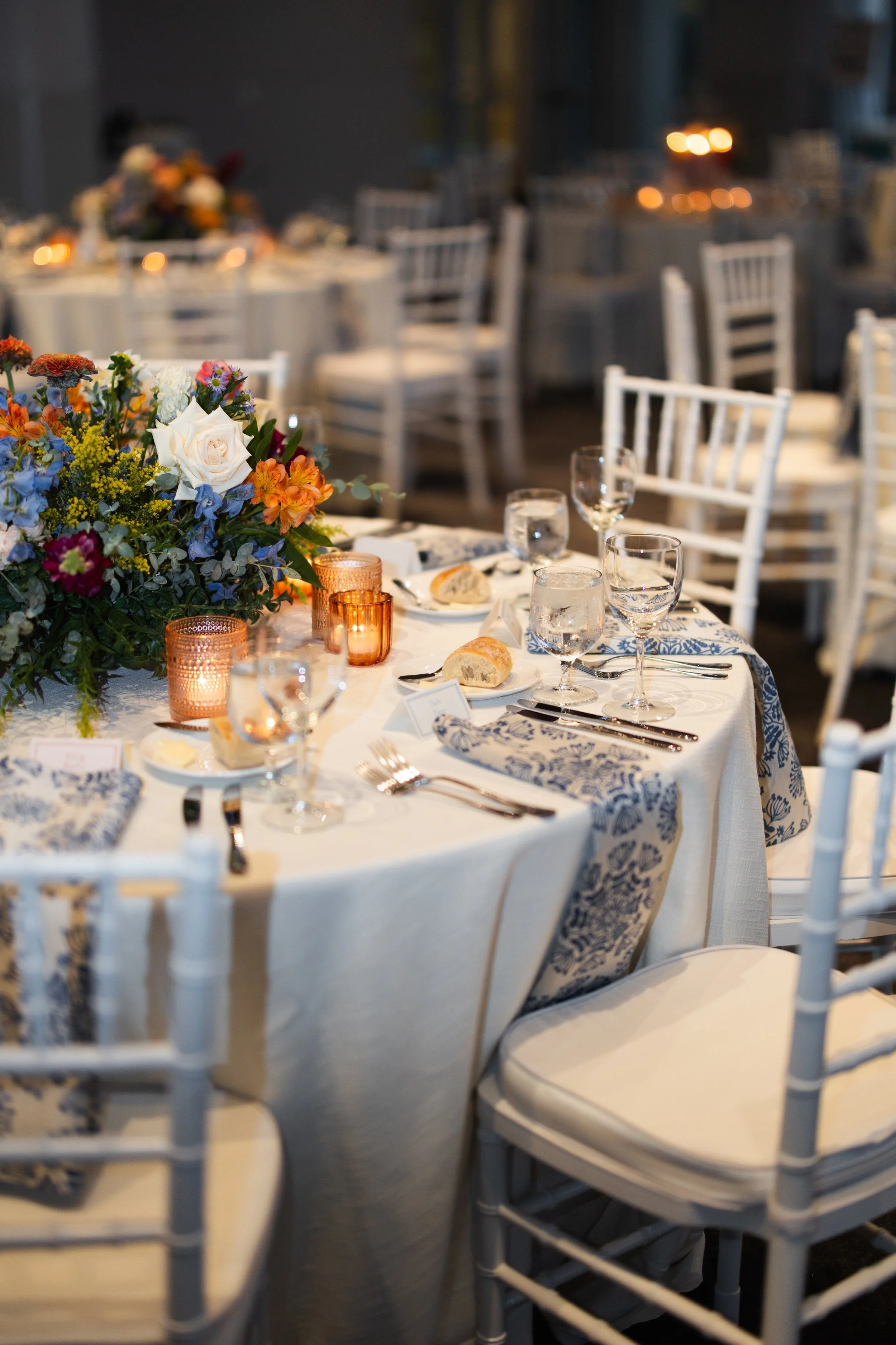 A round banquet table set for a meal with floral centerpiece, candles, glasses, plates, bread, and silverware, surrounded by white chairs in a softly lit indoor setting.