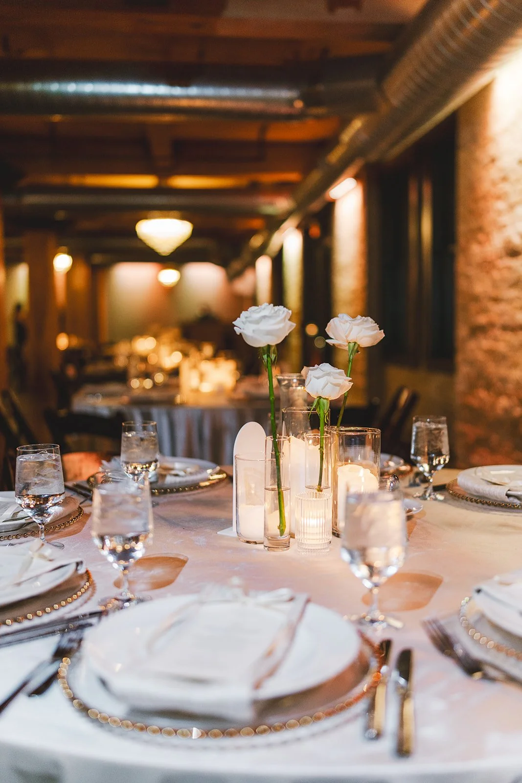 Elegant dining table set with white roses, candles, glassware, white plates, and silverware in a warmly lit restaurant or event space.