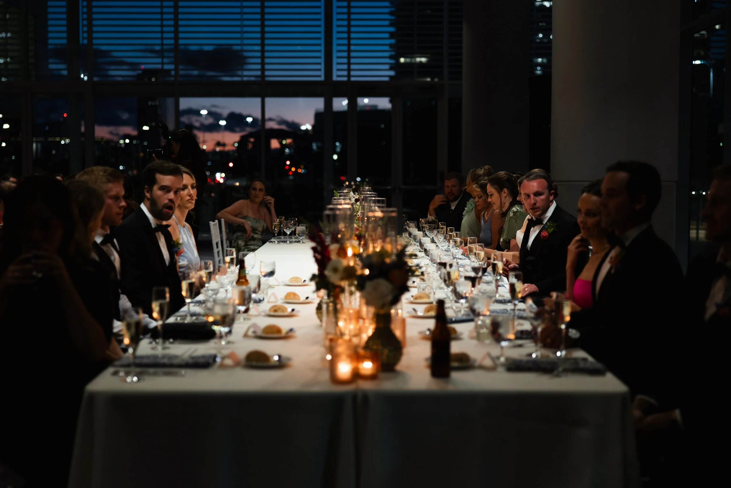 People dressed in formal attire sitting at a long dinner table with candles and floral centerpieces, in a modern venue with large windows showing a sunset sky.