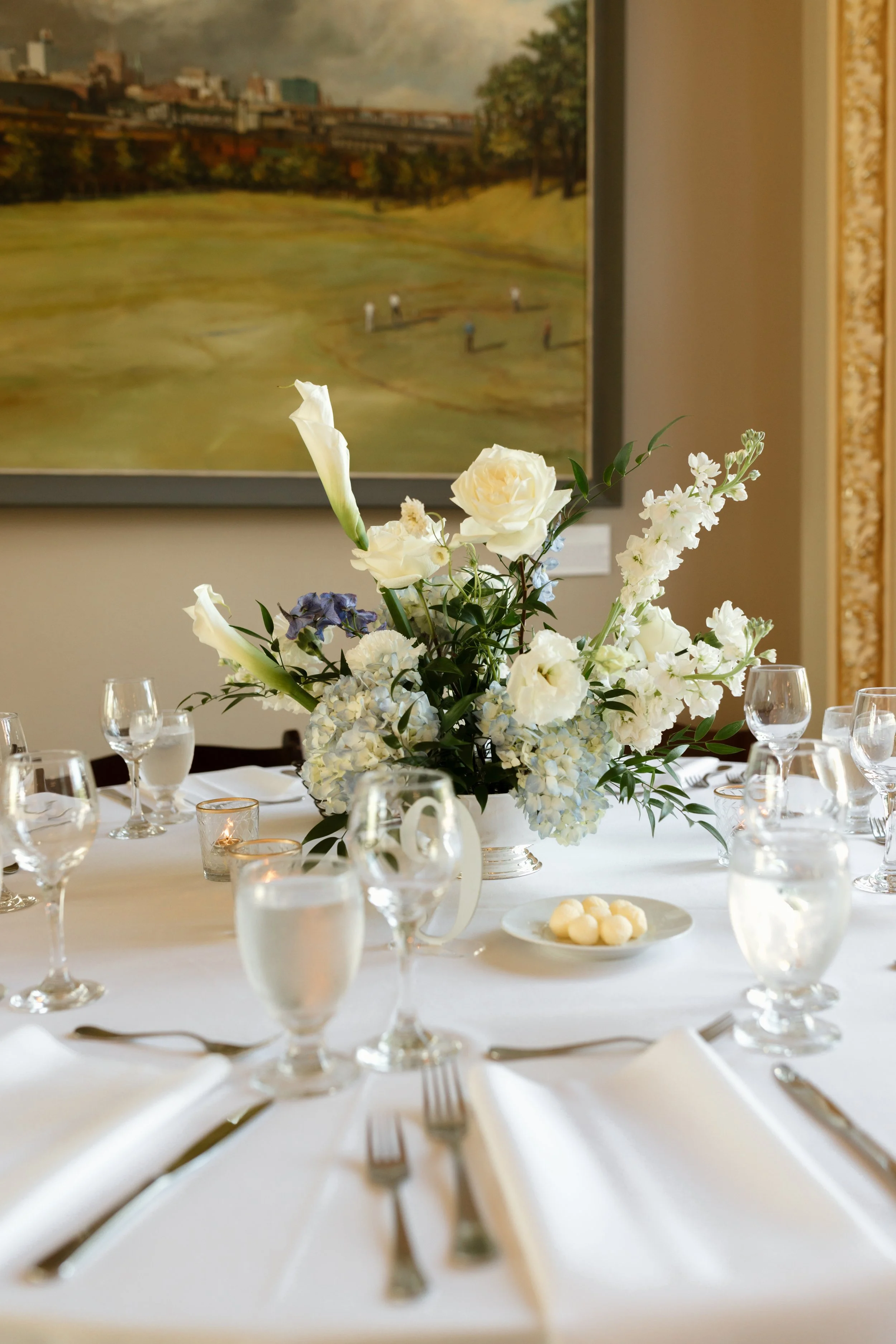 Elegant dining table setting with a large floral centerpiece featuring white roses, calla lilies, and greenery, surrounded by wine glasses, water glasses, and neatly folded white napkins, with candles and a blurred painting of a cityscape in the background.