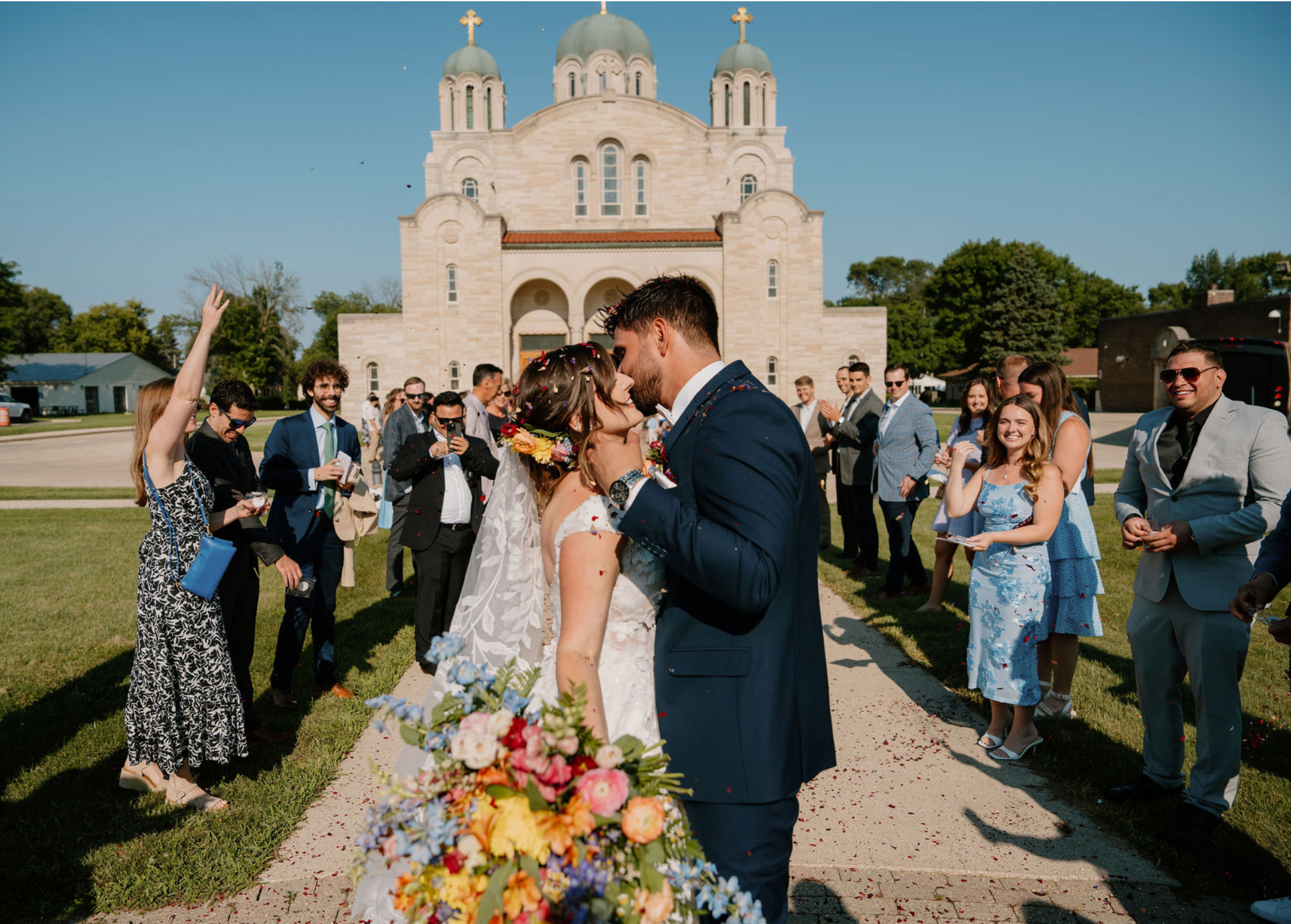 A newlywed couple kissing outdoors in front of a church with guests celebrating and a man holding a large bouquet of colorful flowers in the foreground.