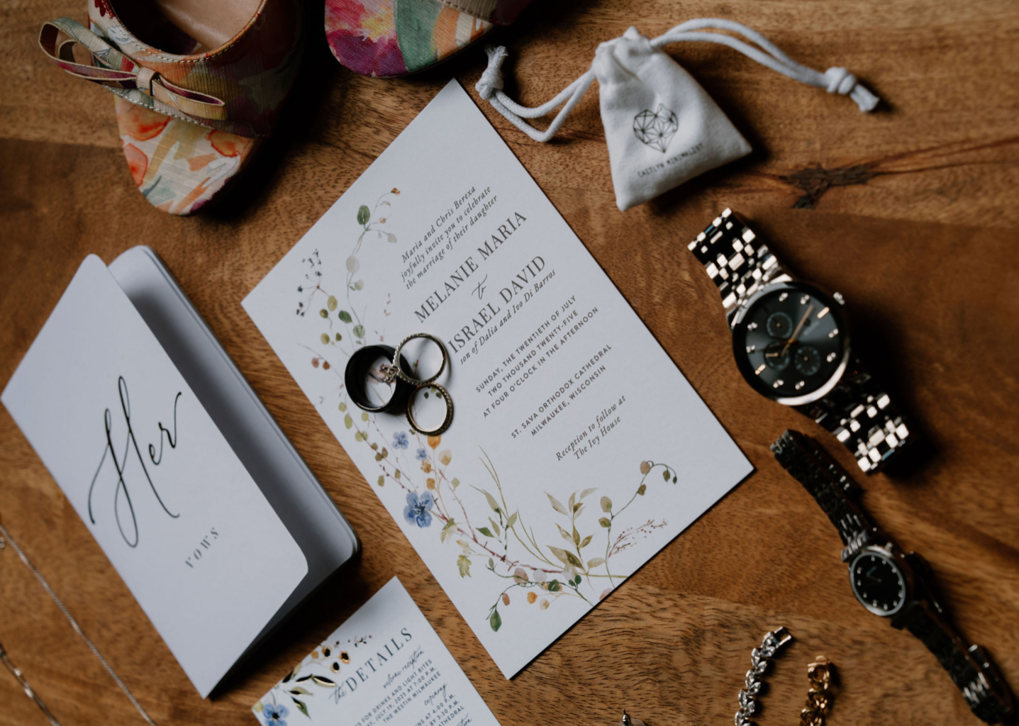 Wedding details: invitation with floral design, rings, watches, and gift box on wooden table.