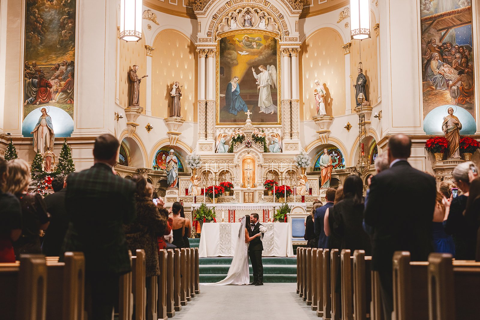 A wedding ceremony inside a church with a bride and groom embracing at the altar, surrounded by guests. The church has ornate religious artwork, statues, and poinsettia plants, indicating it might be during the Christmas season.