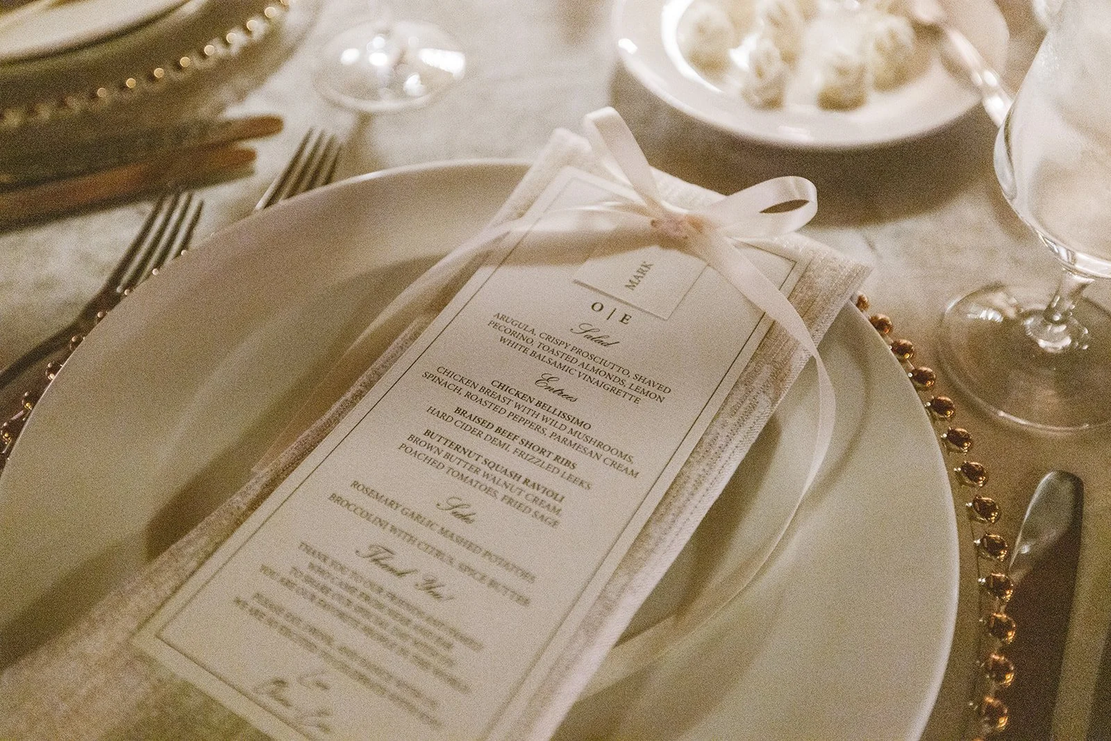 Elegant place setting with a printed menu tied with a ribbon on a napkin, white plate, silverware, a wine glass, and a small plate with food, set on a table with gold bead border.