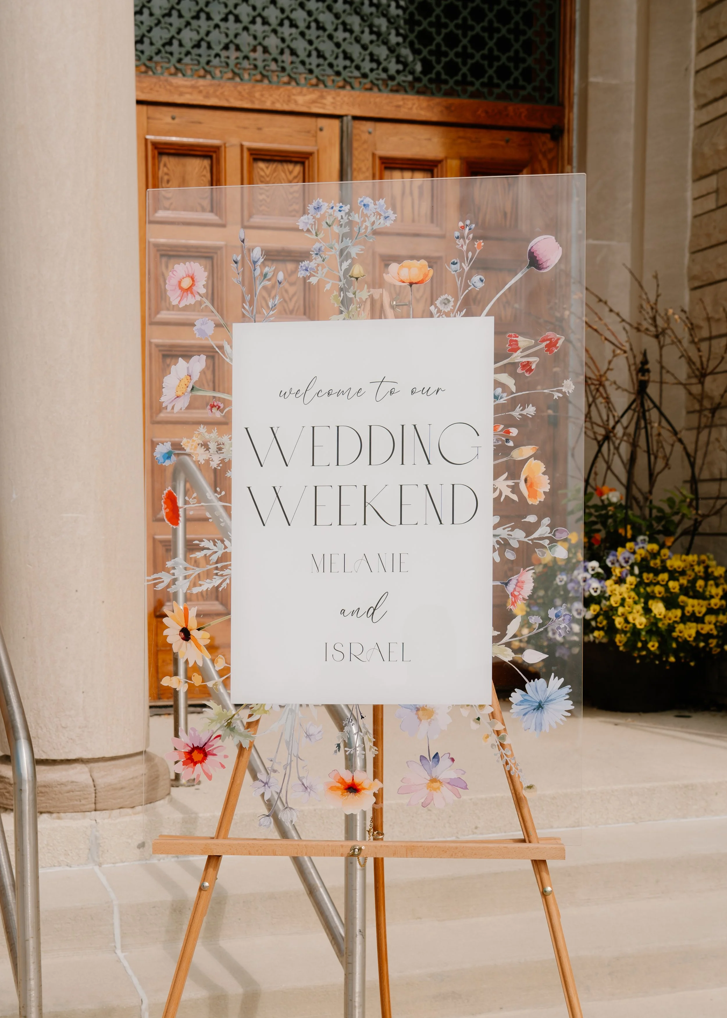 Wedding weekend sign with floral designs on a stand at the entrance of a building, with a wooden door and flowers in the background.
