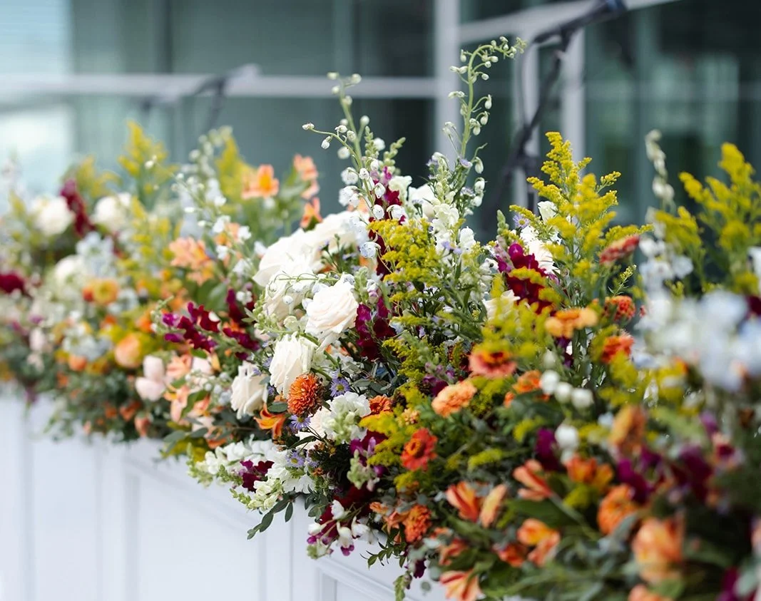 A large colorful bouquet of various flowers including white roses, orange, yellow, and purple blooms, arranged on a white stand or ledge.